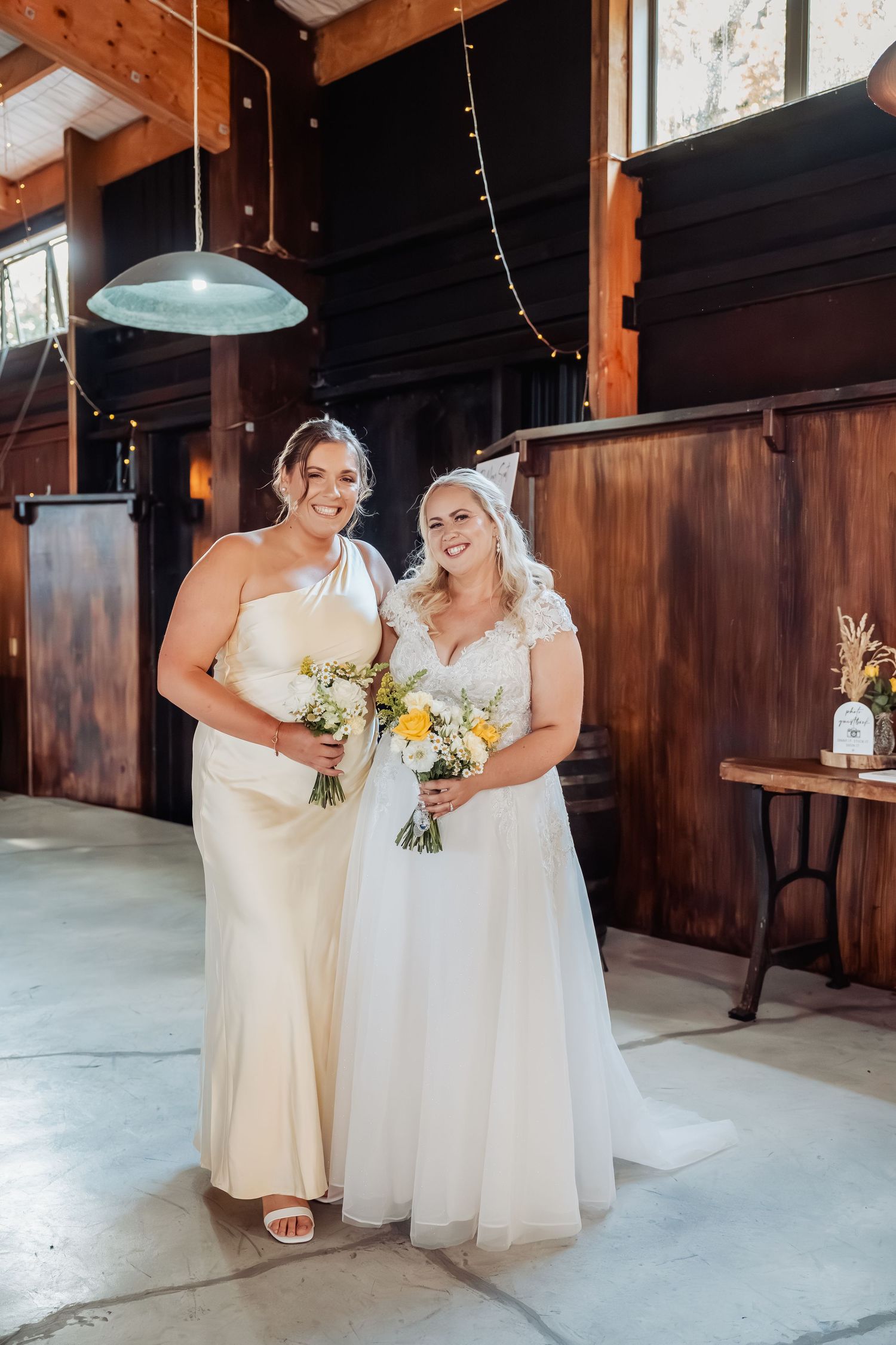 Bride in white tulle gown and bridesmaid in ivory one-shoulder dress posing together in a rustic barn venue.