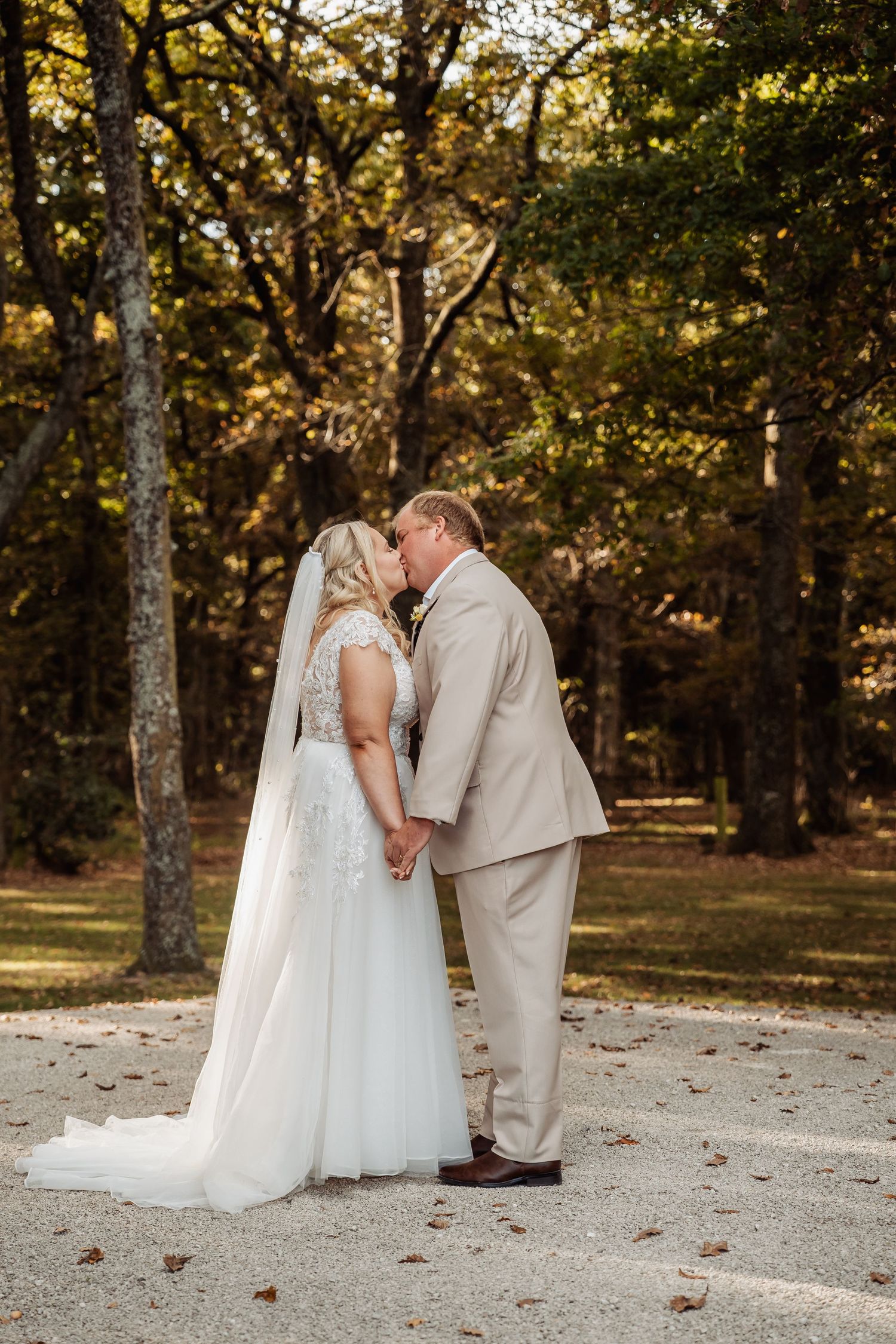 Bride and groom share a kiss while holding hands on a gravel path surrounded by autumn trees.