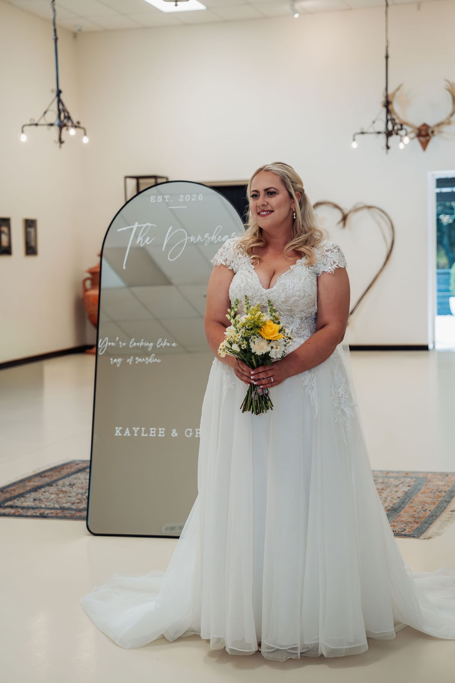 A radiant bride in a white lace wedding gown holds a yellow bouquet beside a personalized wedding mirror sign.