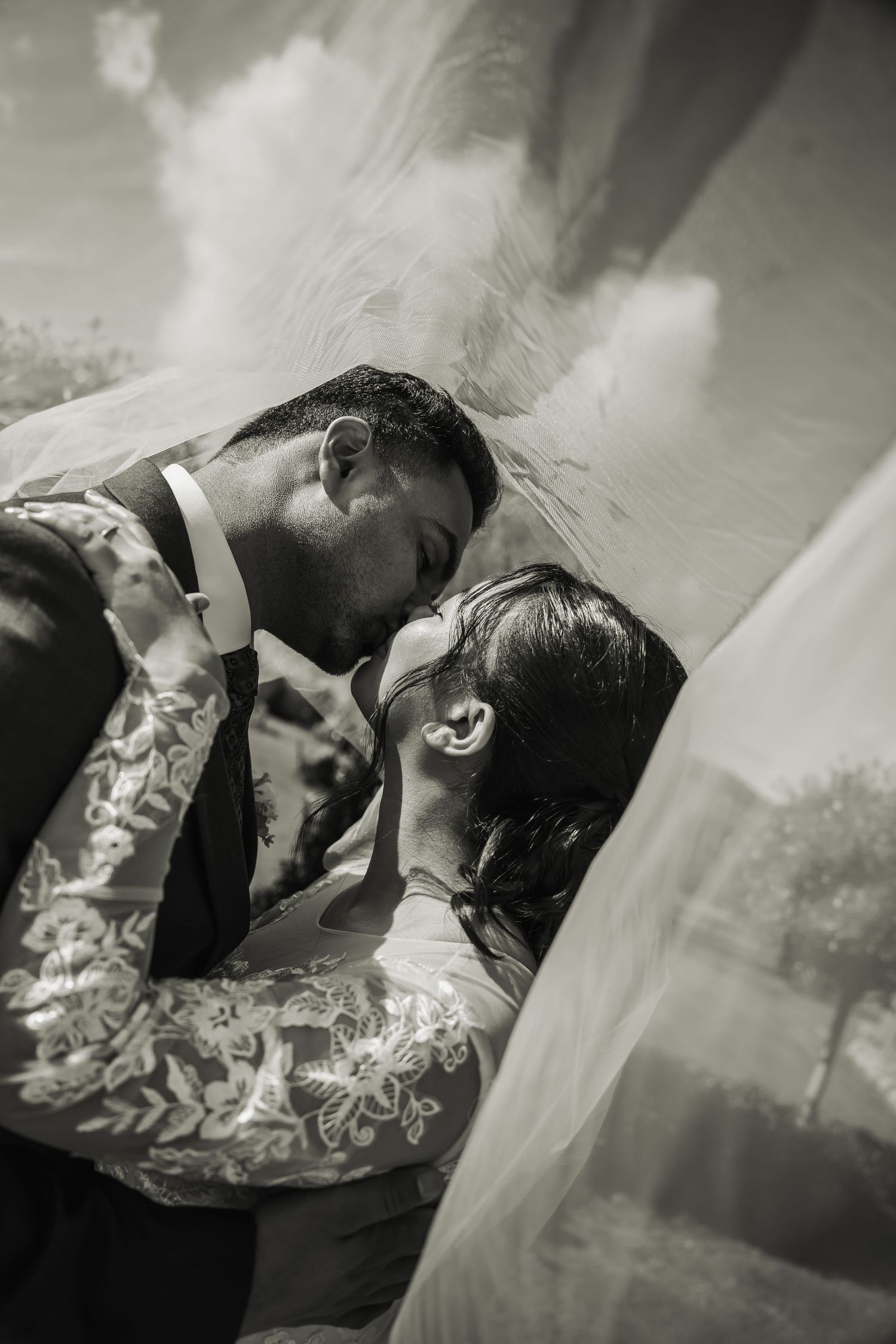 Black and white romantic close-up of bride and groom sharing a kiss beneath flowing wedding veil.