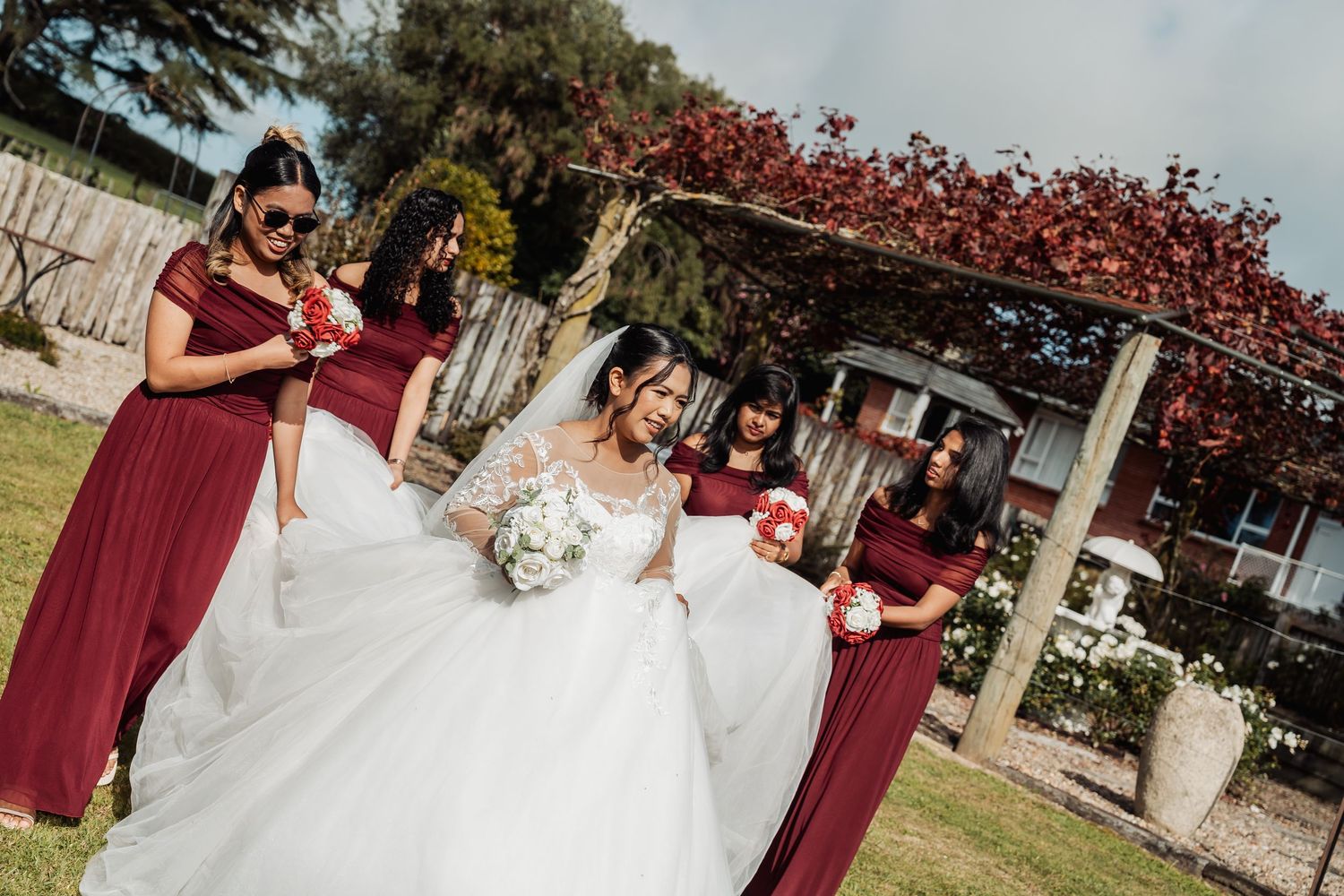 Bride in white lace gown with bridesmaids in burgundy dresses holding bouquets in sunny garden.