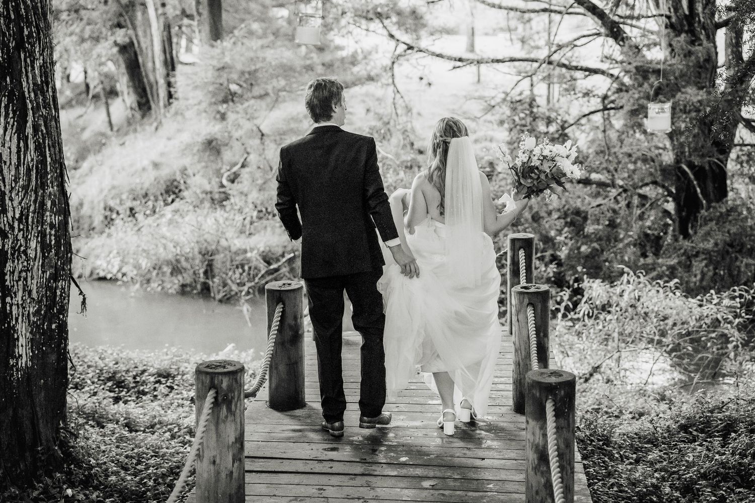 A wedding couple walks together across a rustic wooden bridge in a black and white forest setting.