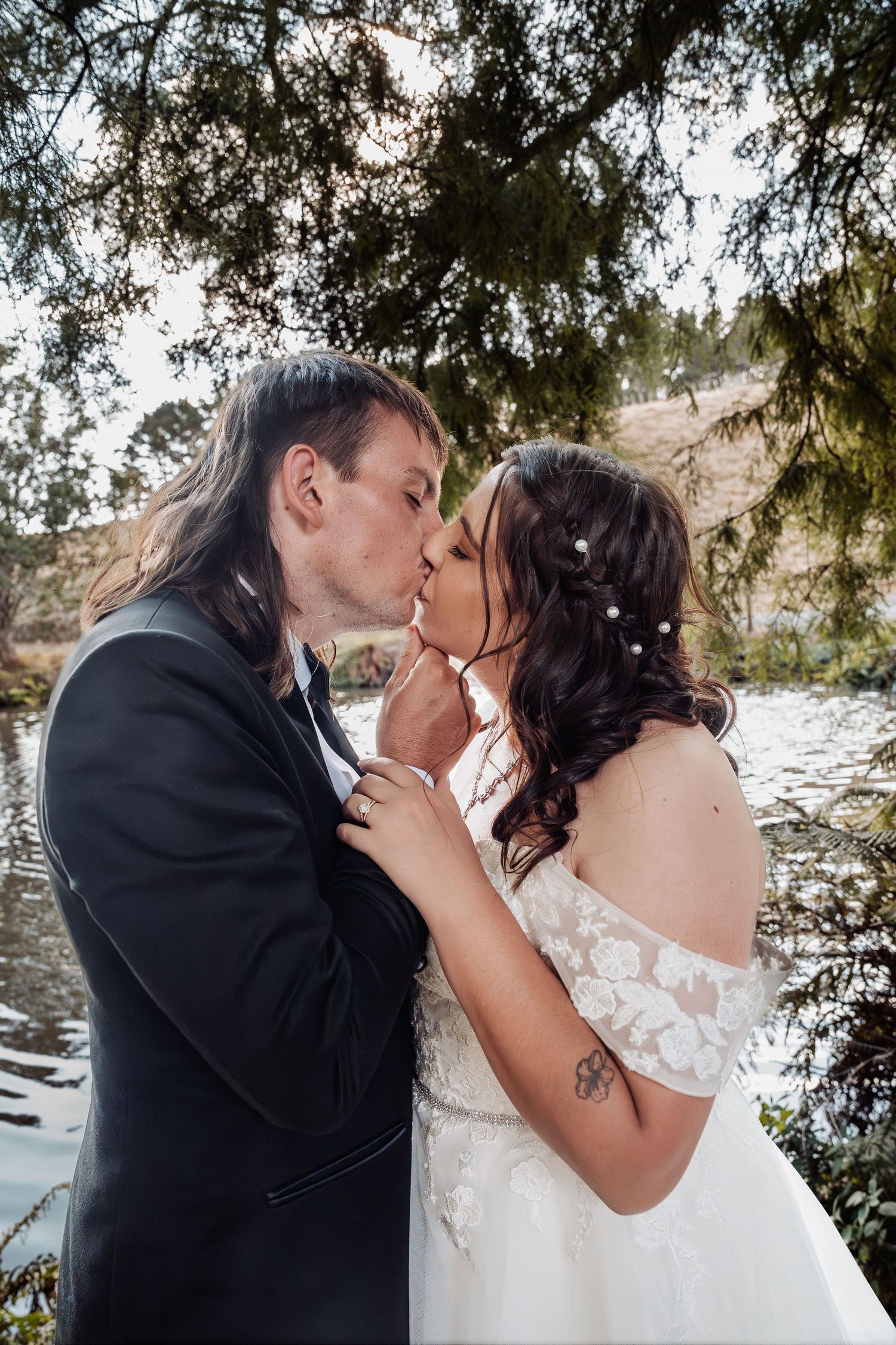 Groom tenderly kisses bride near a serene pond under overhanging trees at their wedding.