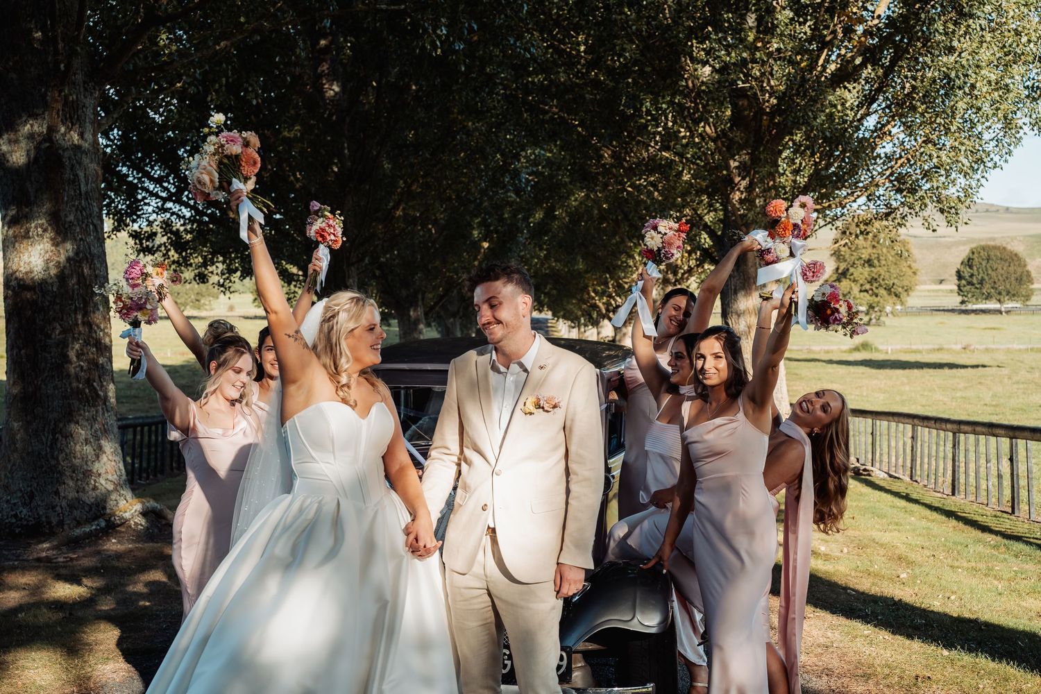 Bride and groom celebrating with wedding party, raising bouquets joyfully under shady trees.