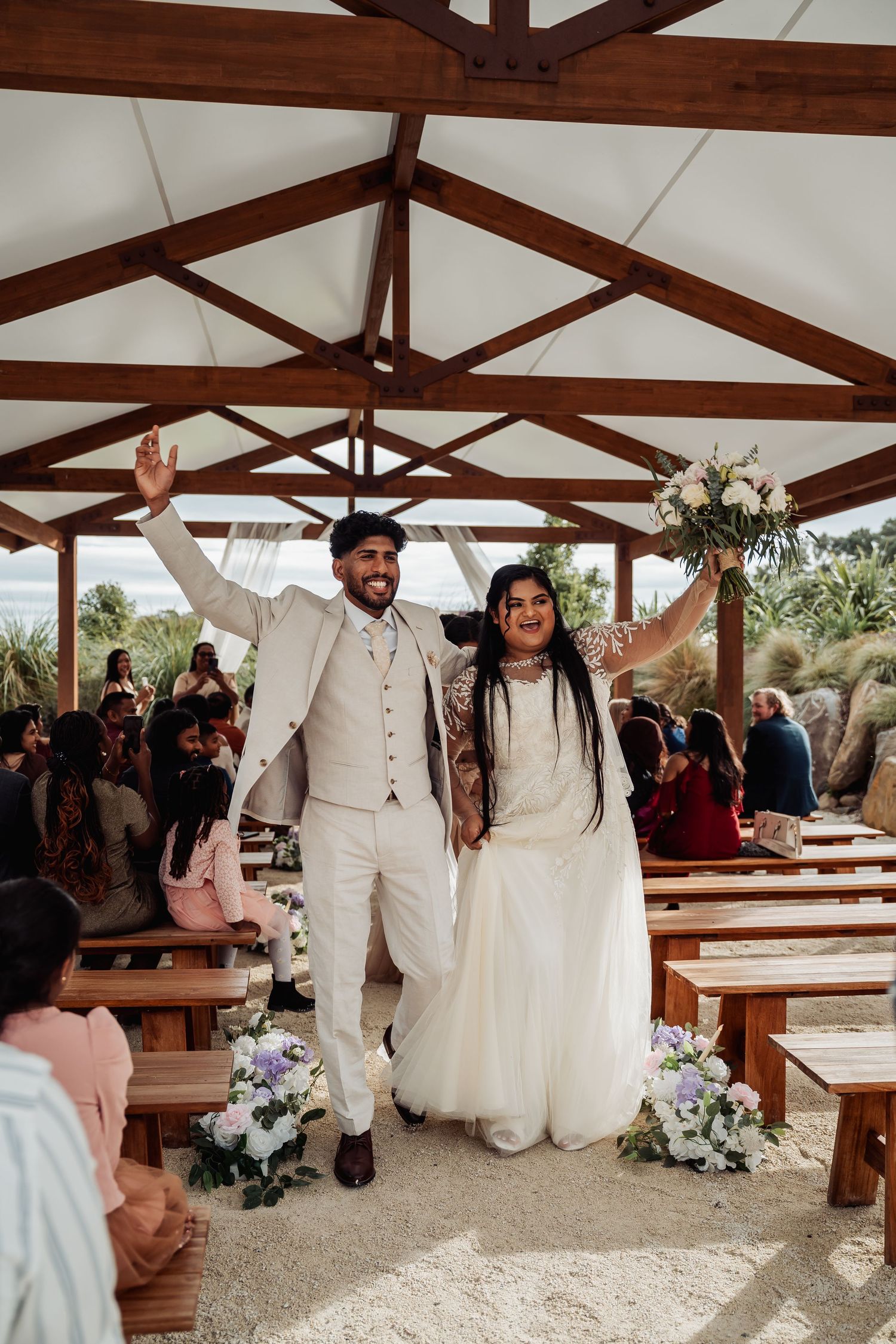 Joyful newlyweds celebrate walking down the aisle at an outdoor wedding ceremony under a wooden pergola.