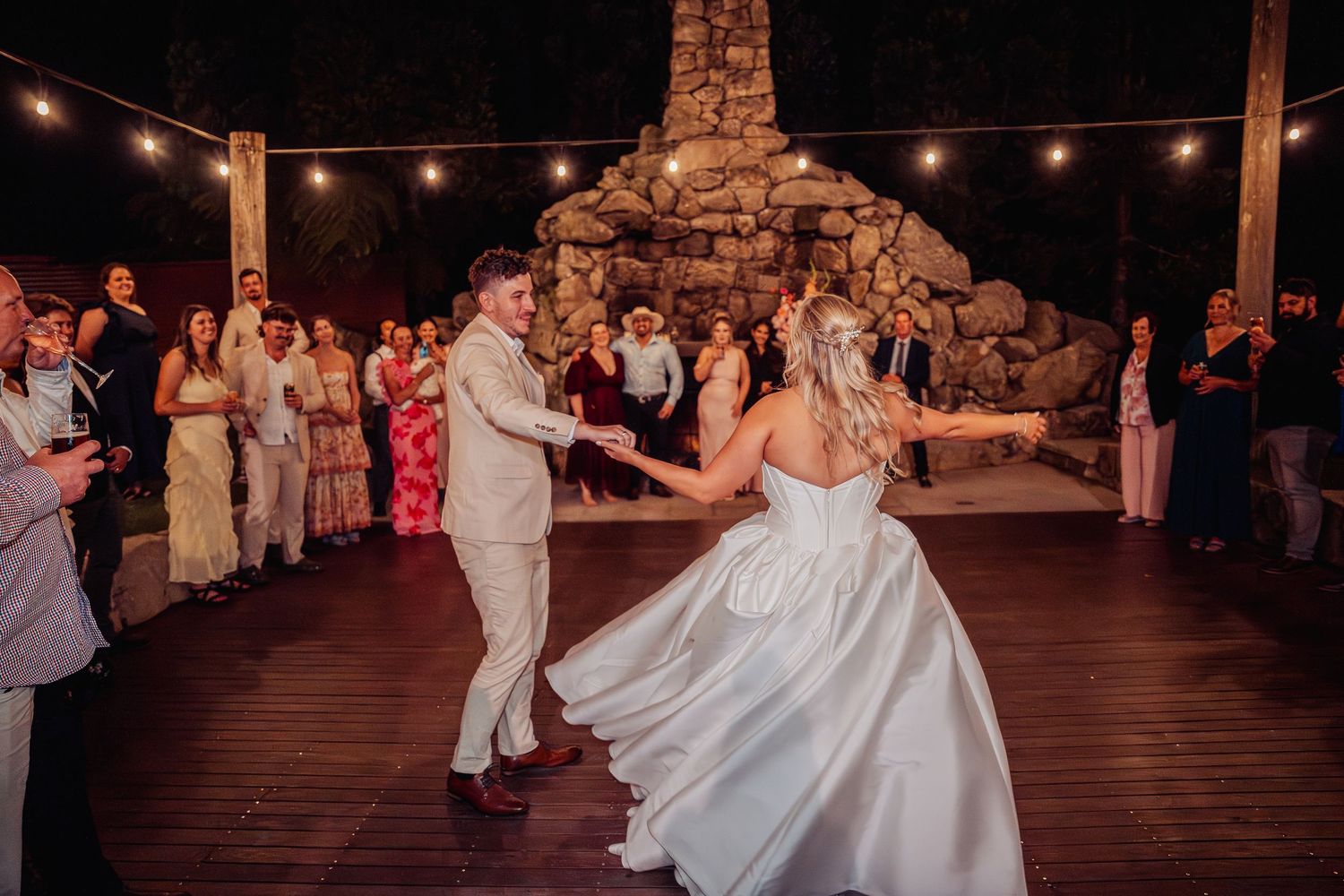 Bride and groom share first dance on outdoor deck with guests watching at night.