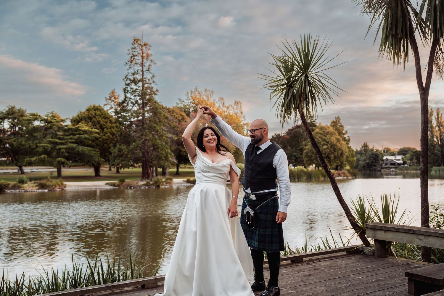 Bride and groom in tartan kilt dancing joyfully on a wooden deck beside a tranquil lake at sunset.