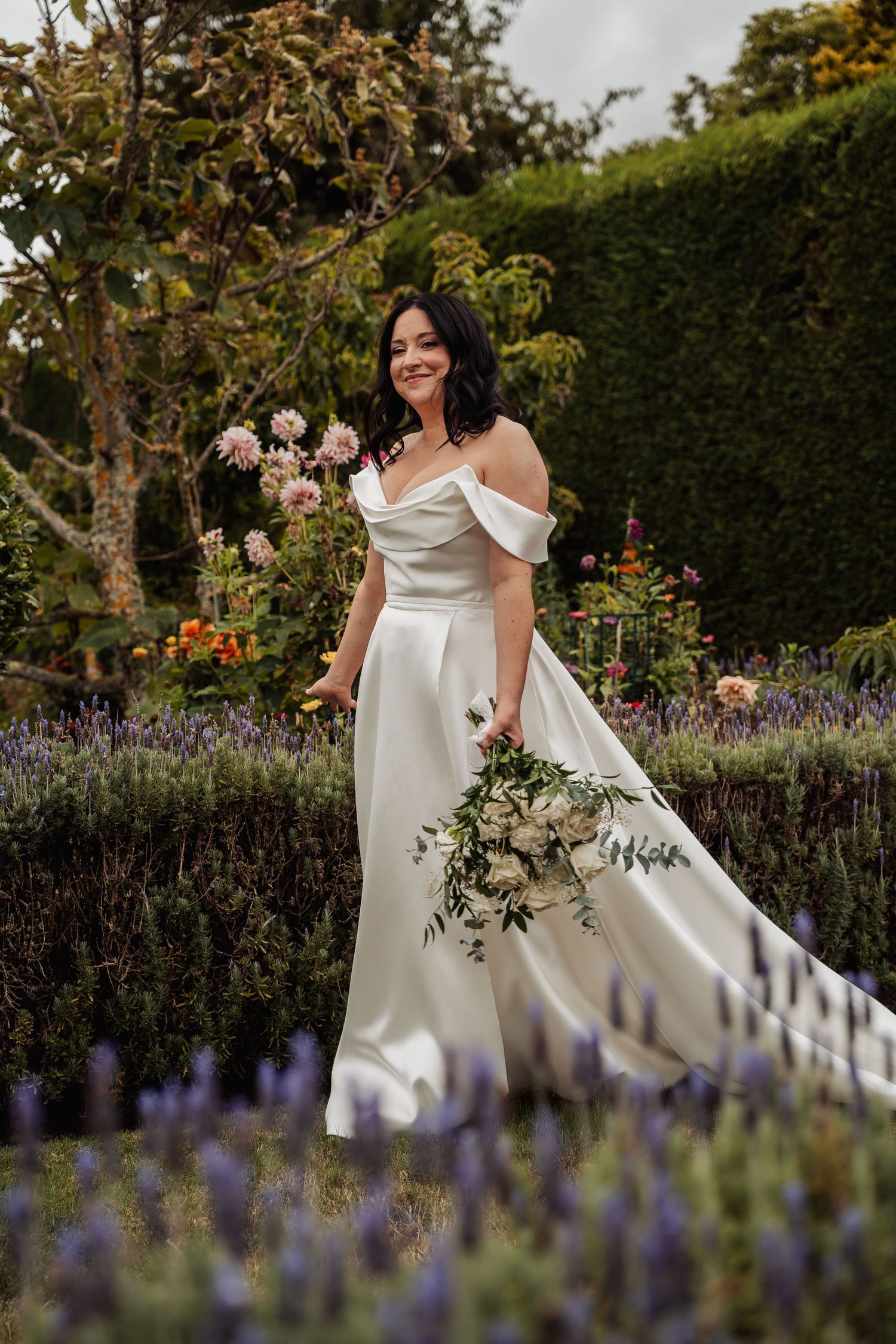 Smiling bride in elegant off-shoulder satin gown holds white bouquet among lavender and flowers in garden.