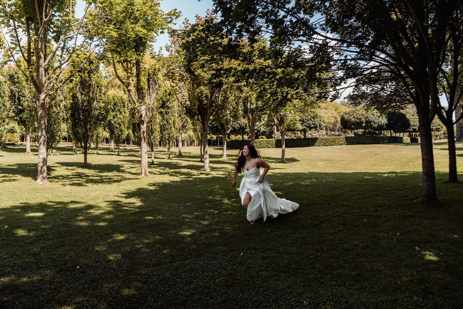 Bride in white gown running through a lush green park surrounded by tall trees on a sunny day.