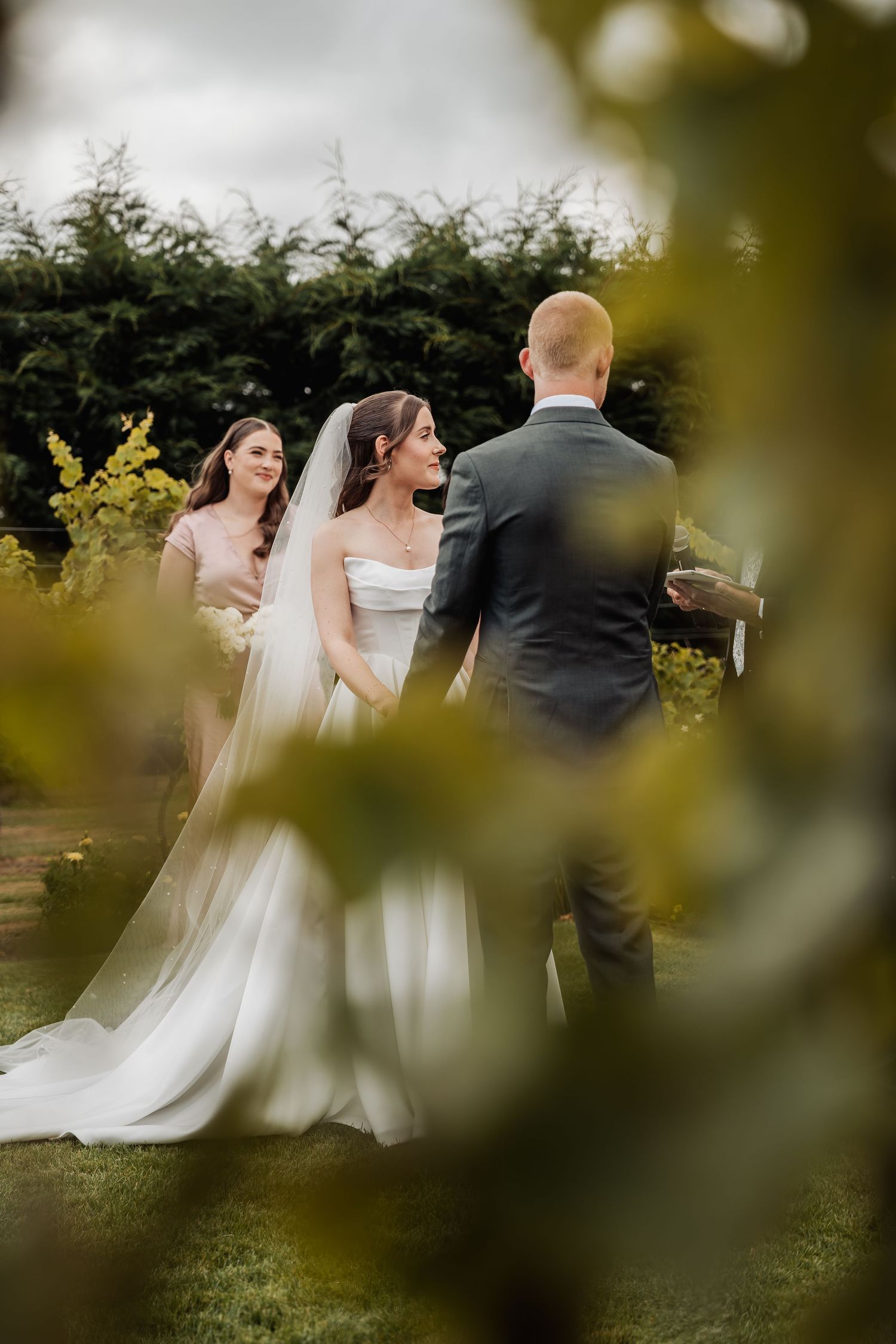 Bride and groom exchange vows in an outdoor garden ceremony, viewed through soft green foliage.