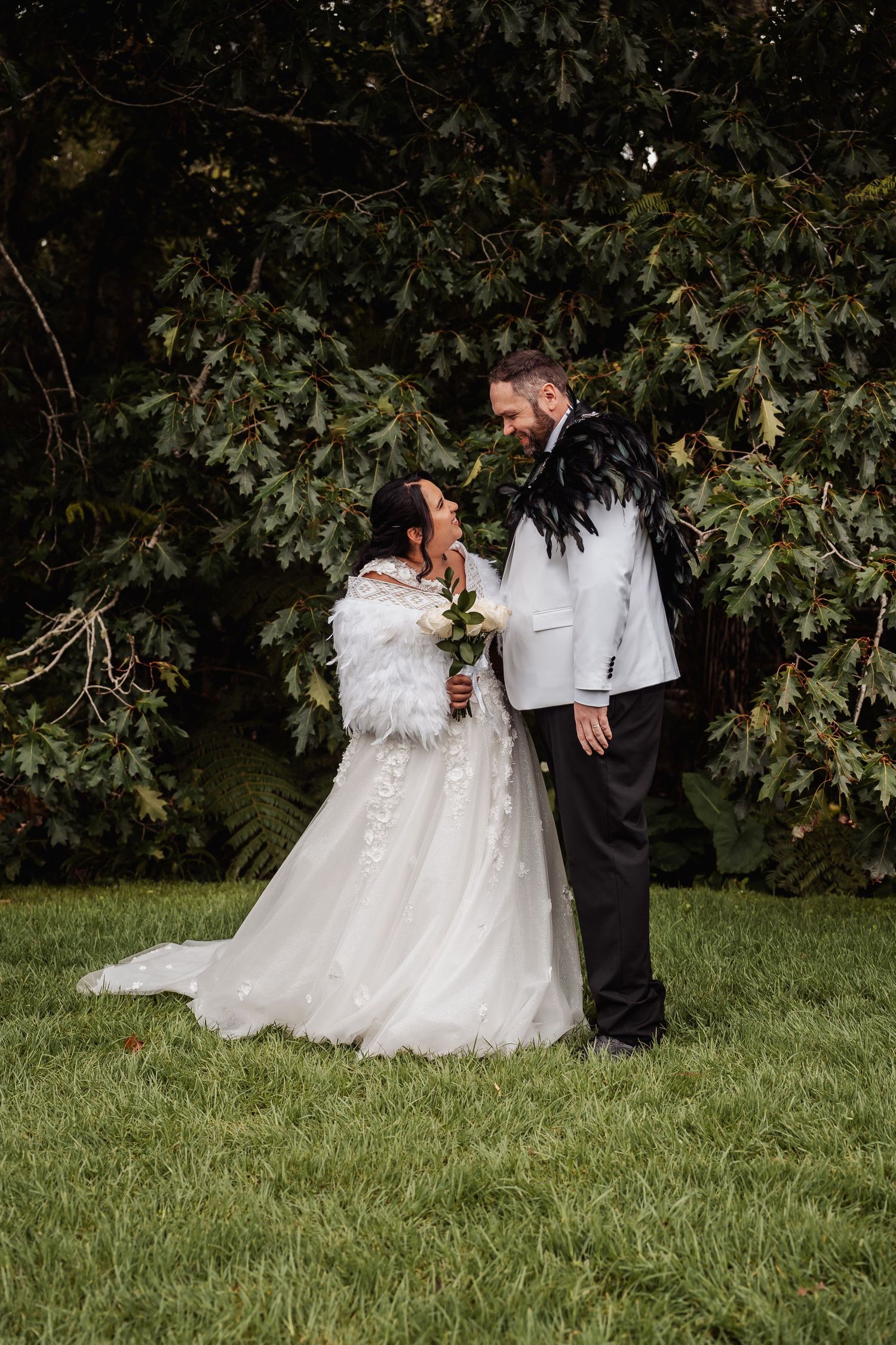 Bride and groom share romantic moment in garden wearing white dress and feathered jacket.
