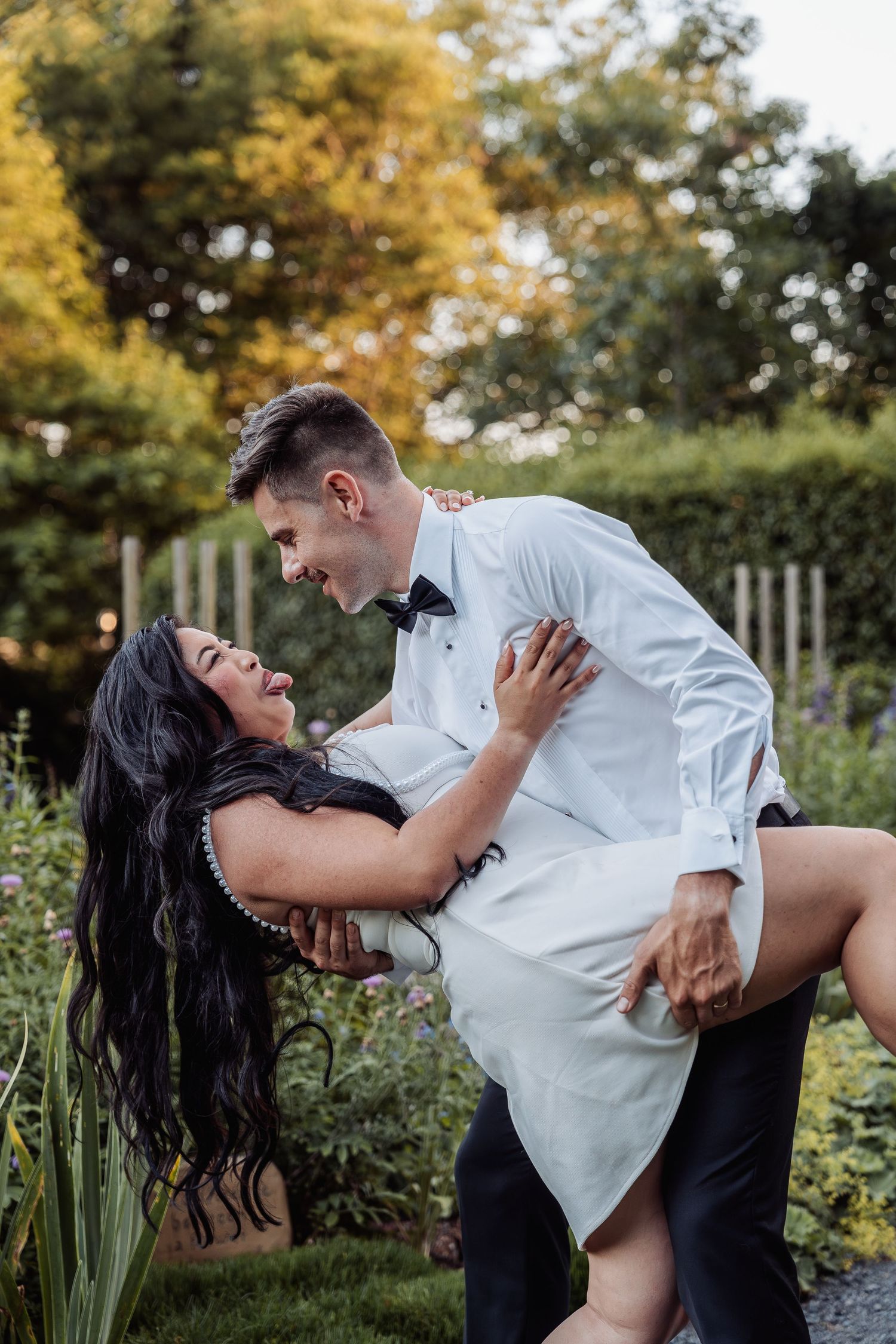 Joyful groom lifts laughing bride in pearl necklace and white dress in lush garden setting.