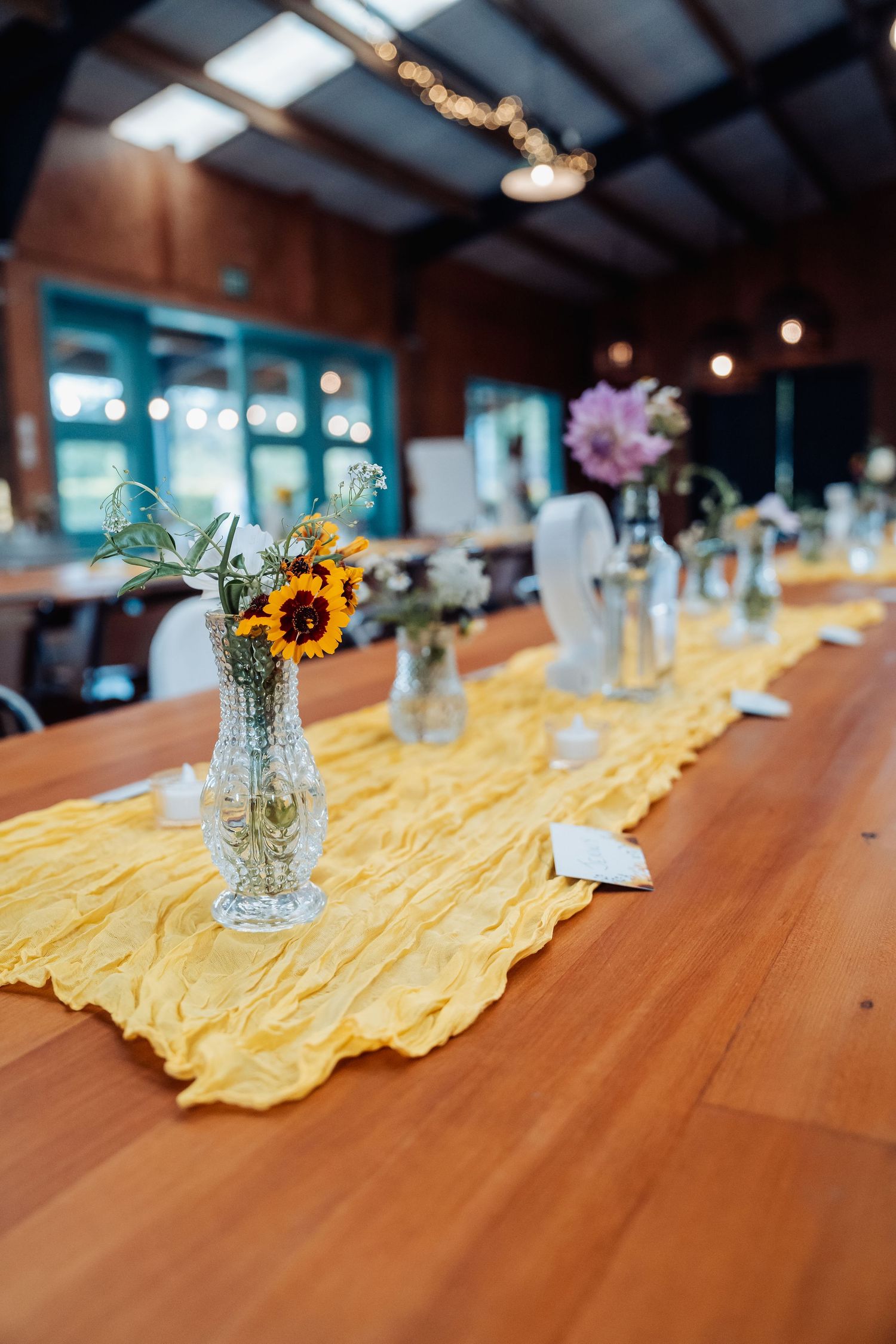 Rustic wedding reception table with yellow runner and sunflower centerpieces in crystal vases.