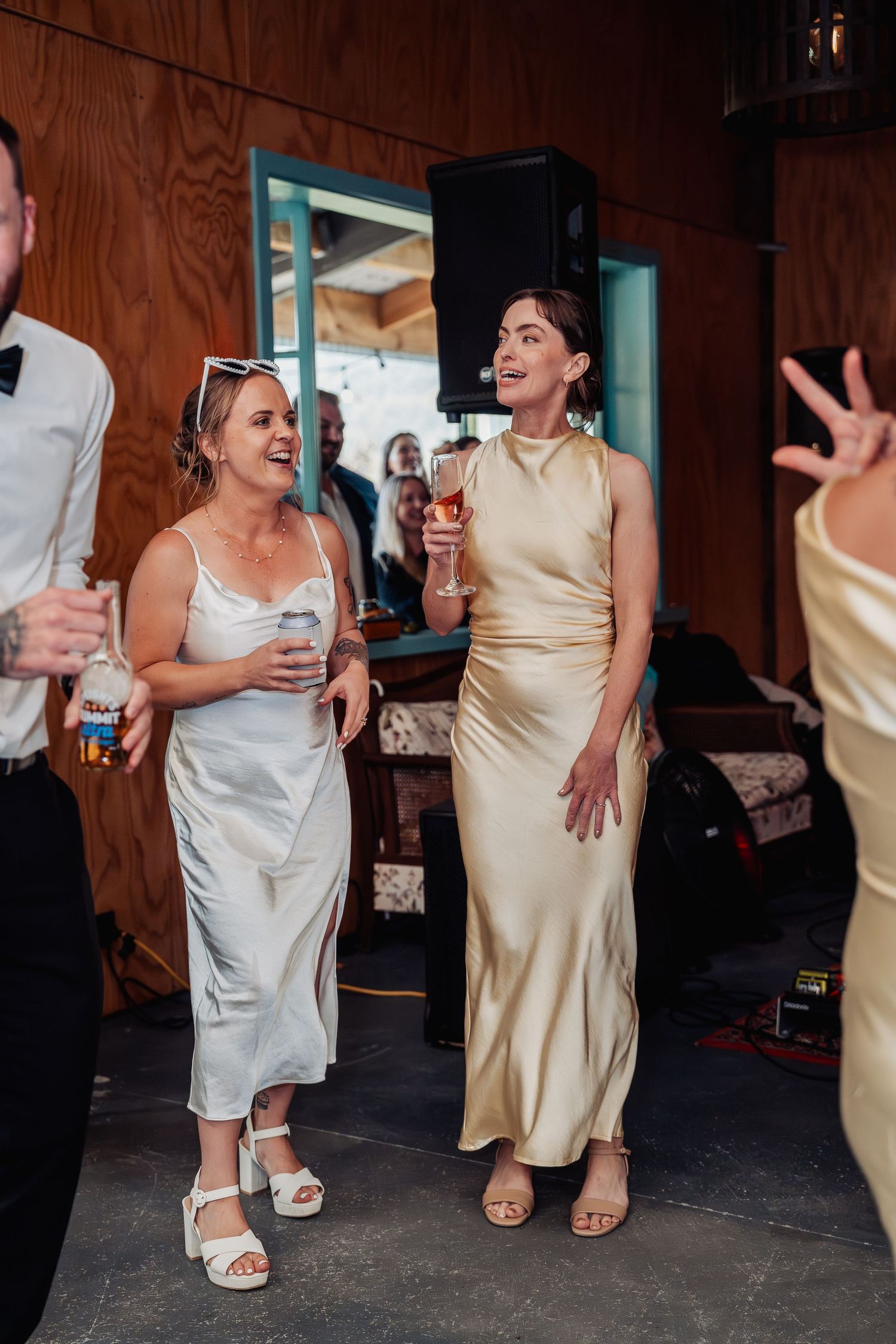 Two women in elegant dresses laughing and dancing at a wedding reception, one in white and one in gold satin.