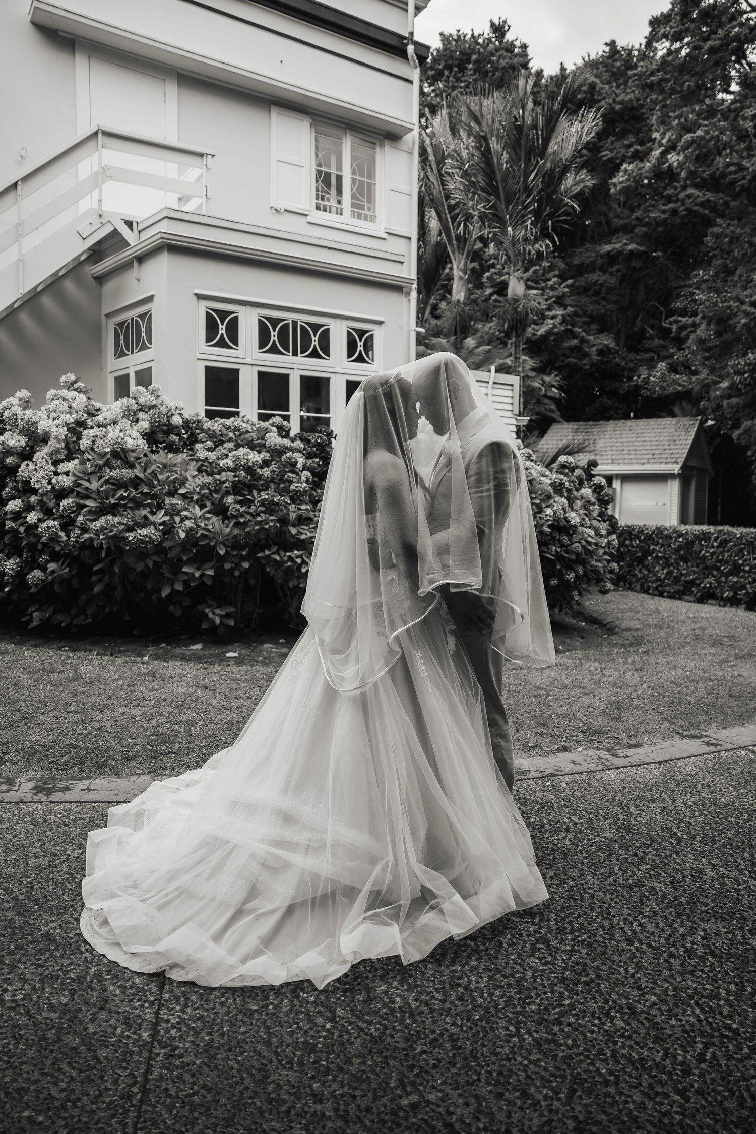 Black and white photo of bride and groom kissing under flowing veil outside elegant venue.