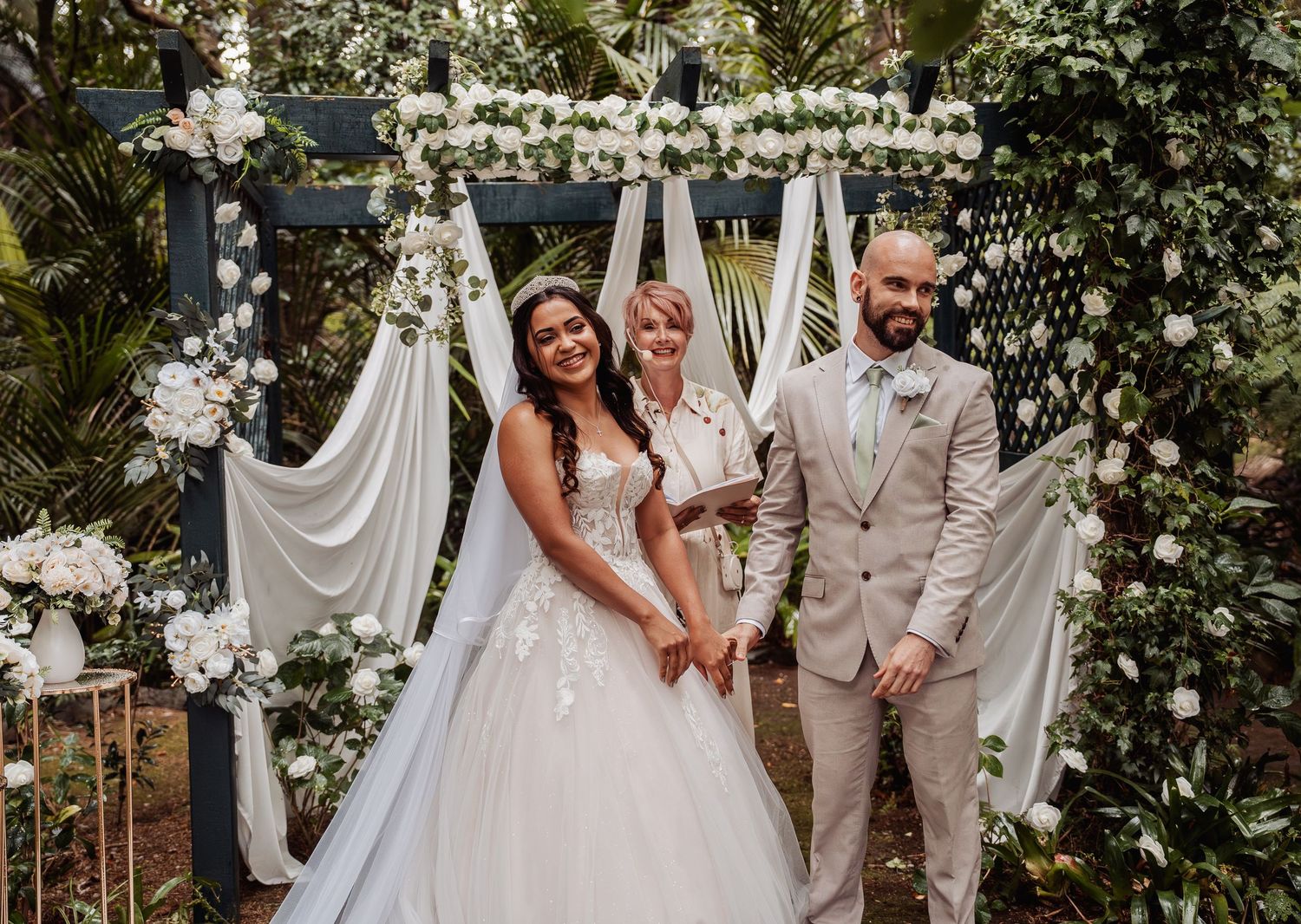 Bride and groom stand with officiant under floral arch with white draping and roses at outdoor wedding ceremony.