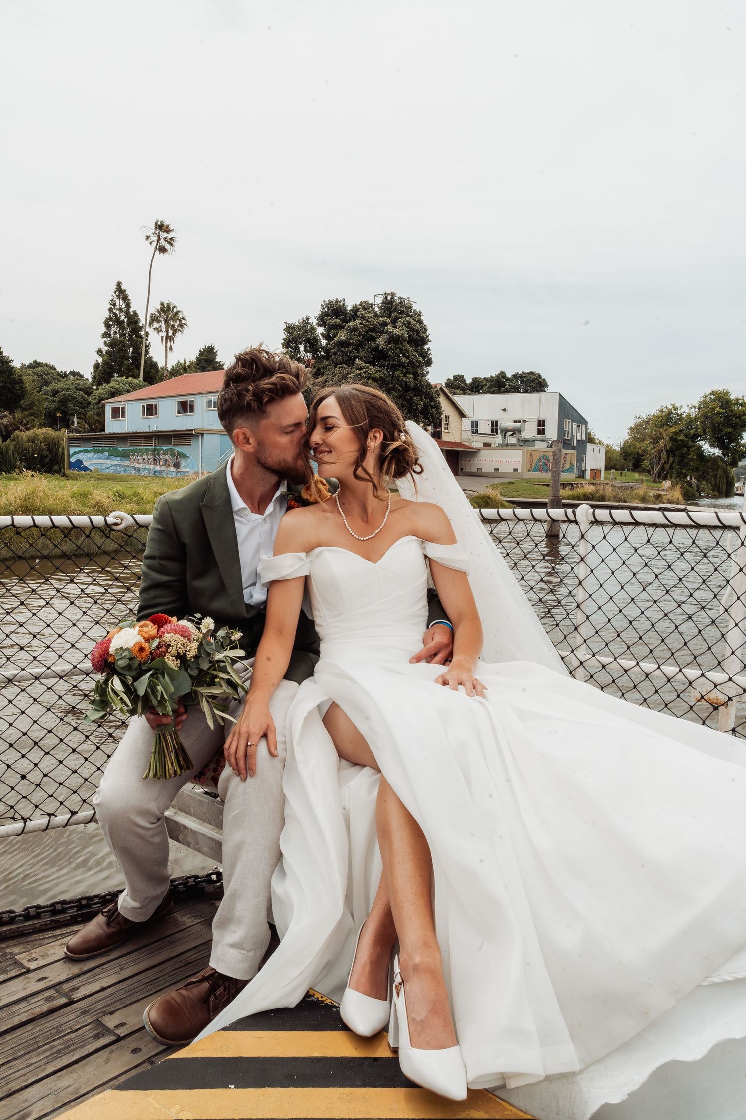 Bride and groom sitting together on a dock, sharing a tender kiss with a bouquet.
