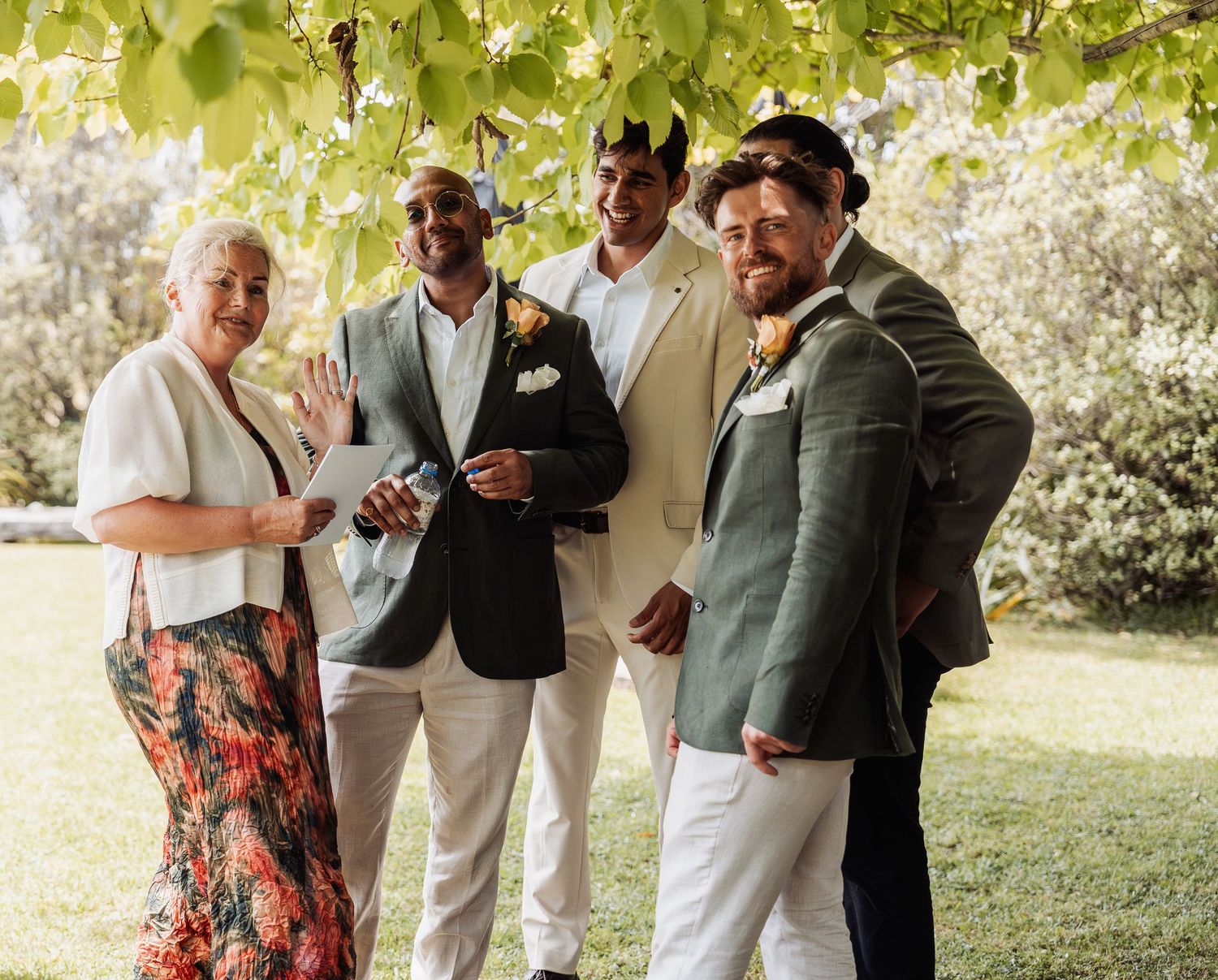 Four wedding guests posing and smiling under leafy vines in a sunny outdoor garden setting.