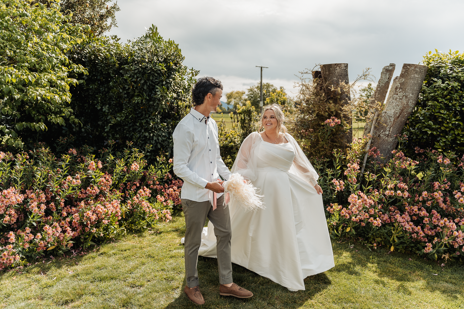 A couple stands together in a manicured garden with pink roses and hedges.