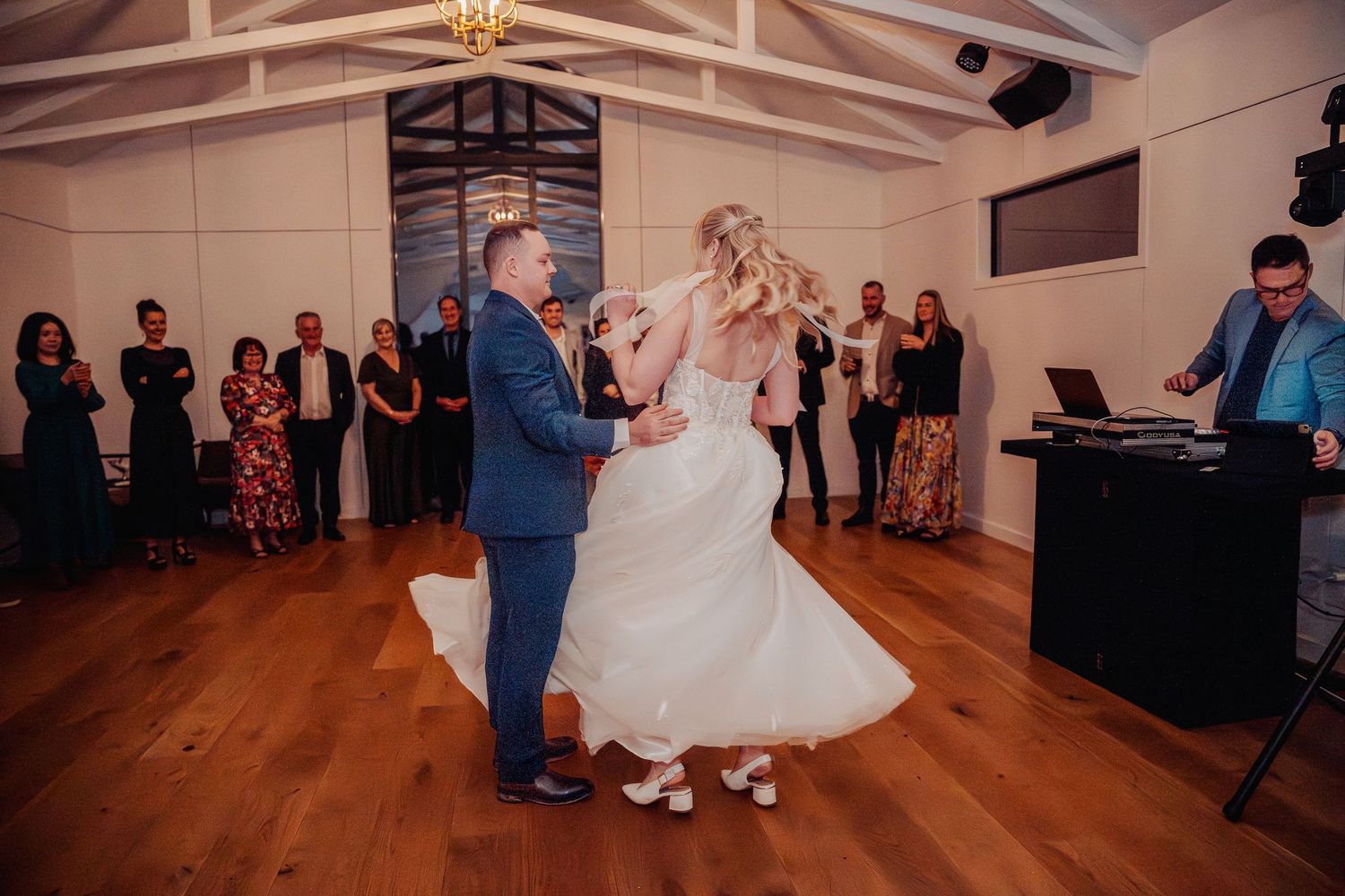 Wedding couple's first dance on wooden floor with guests watching from the sides.