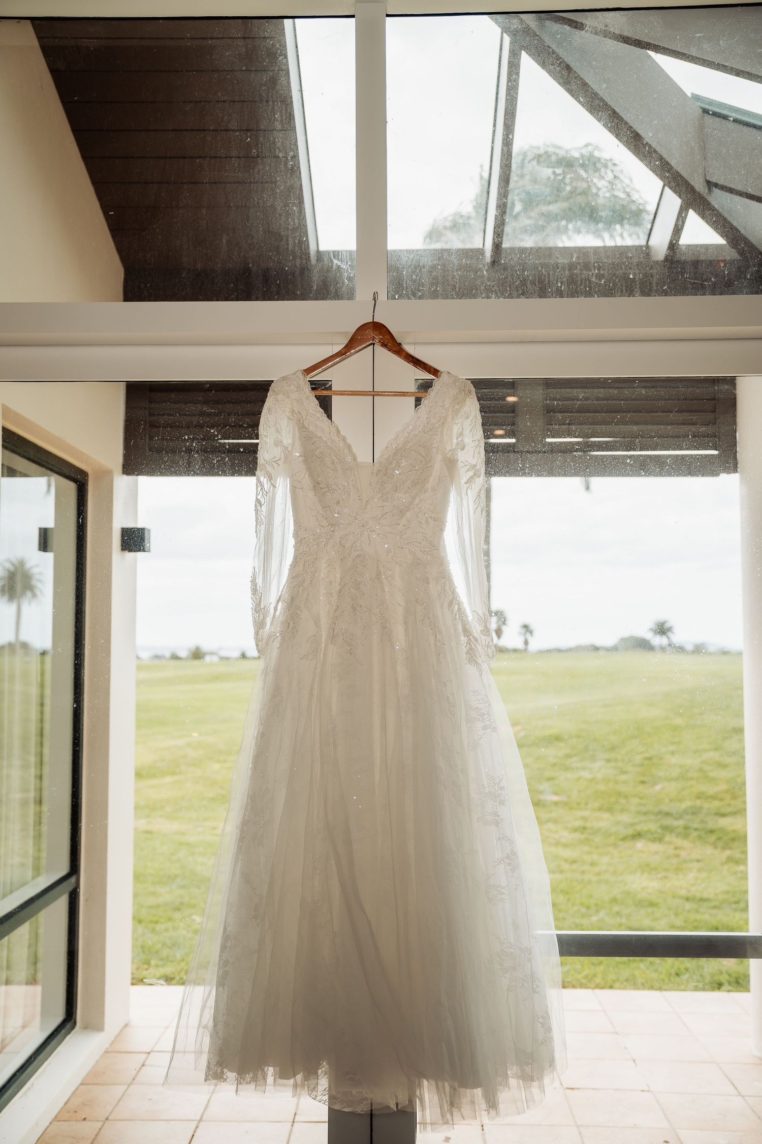 White wedding dress hanging in front of large windows with natural light streaming in and green landscape visible outside.
