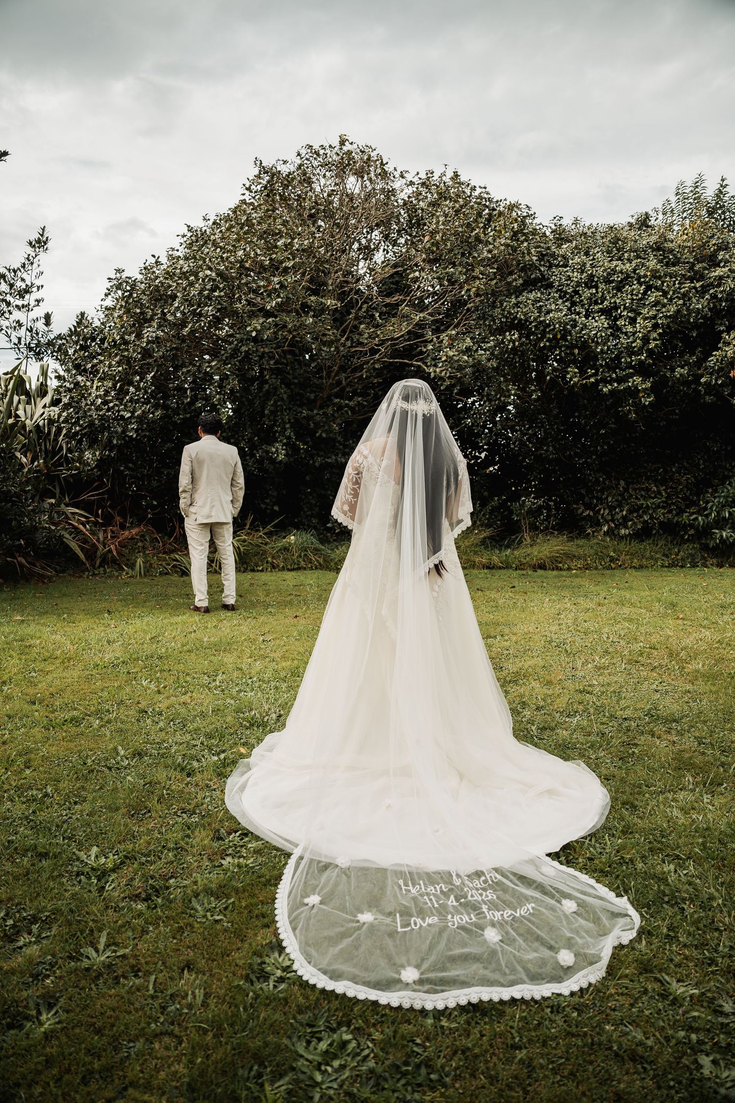 Bride in flowing white gown with long embroidered veil stands facing groom in beige suit on lush green lawn.