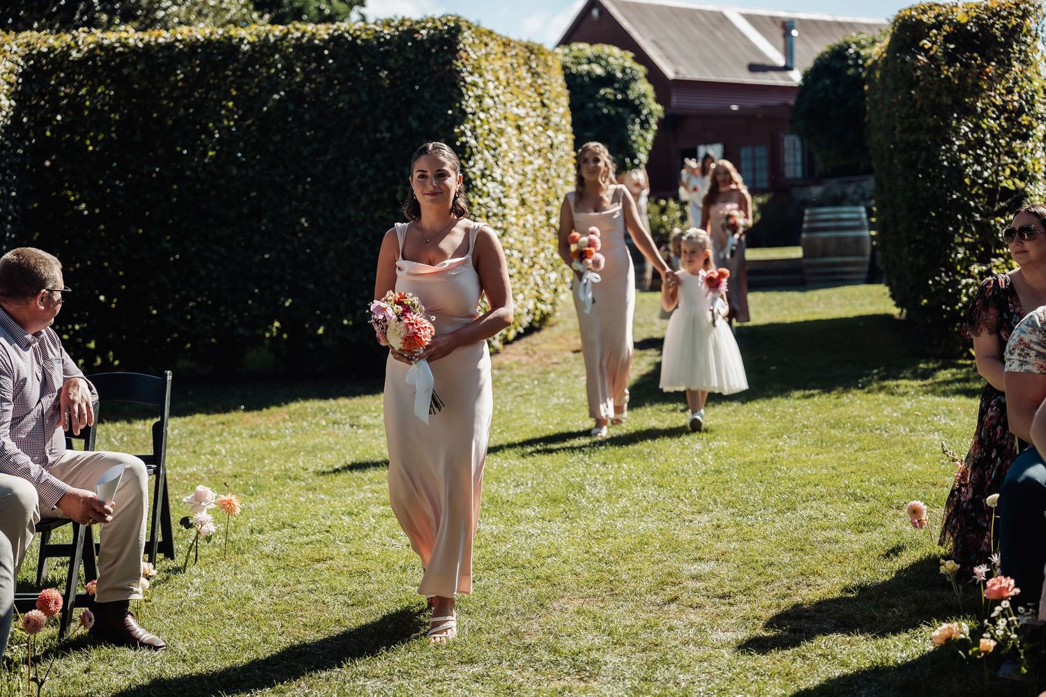 Bridesmaids in blush pink dresses walking down outdoor grass aisle carrying bouquets at a sunny wedding.