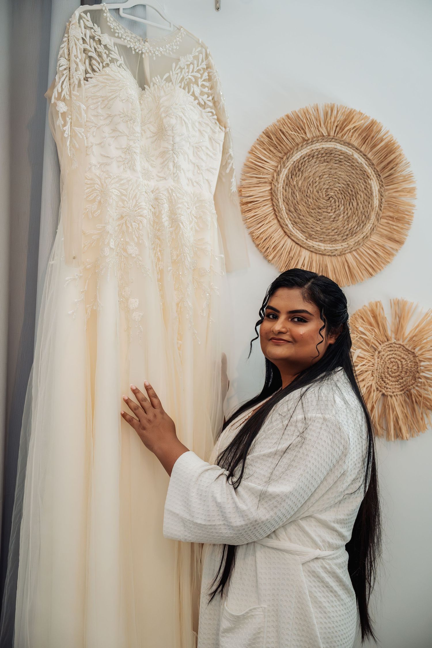 A bride in a white robe admires her lace wedding gown hanging on display with woven wall decor behind her.