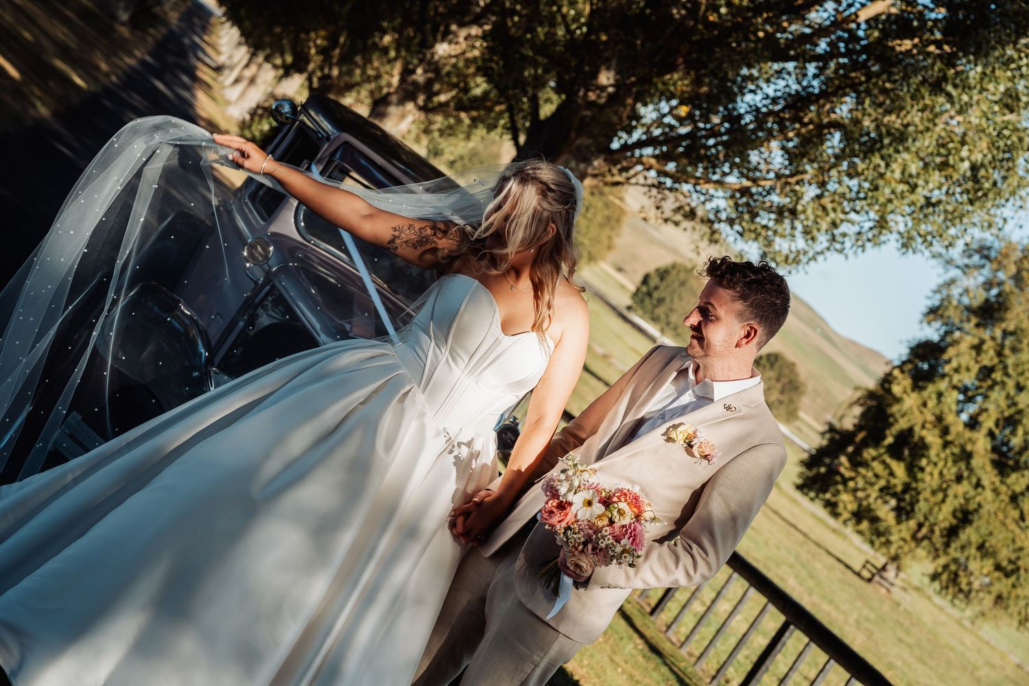 Groom laughing as bride's veil blows in the wind beside a vintage car on their wedding day.