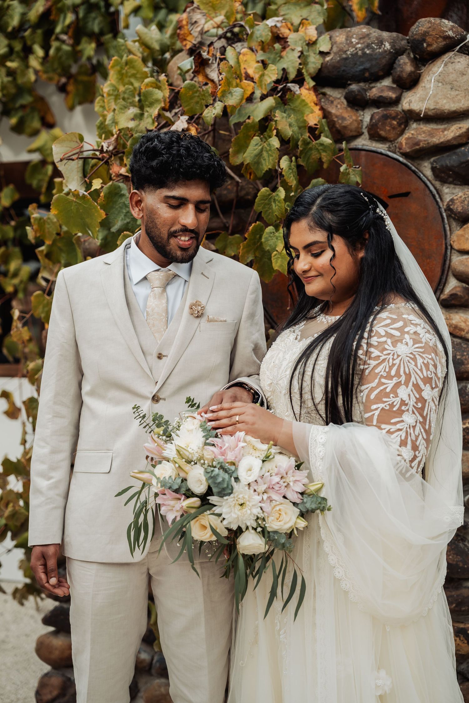 Bride and groom in beige suit admire pastel floral bouquet together against stone wall with foliage.