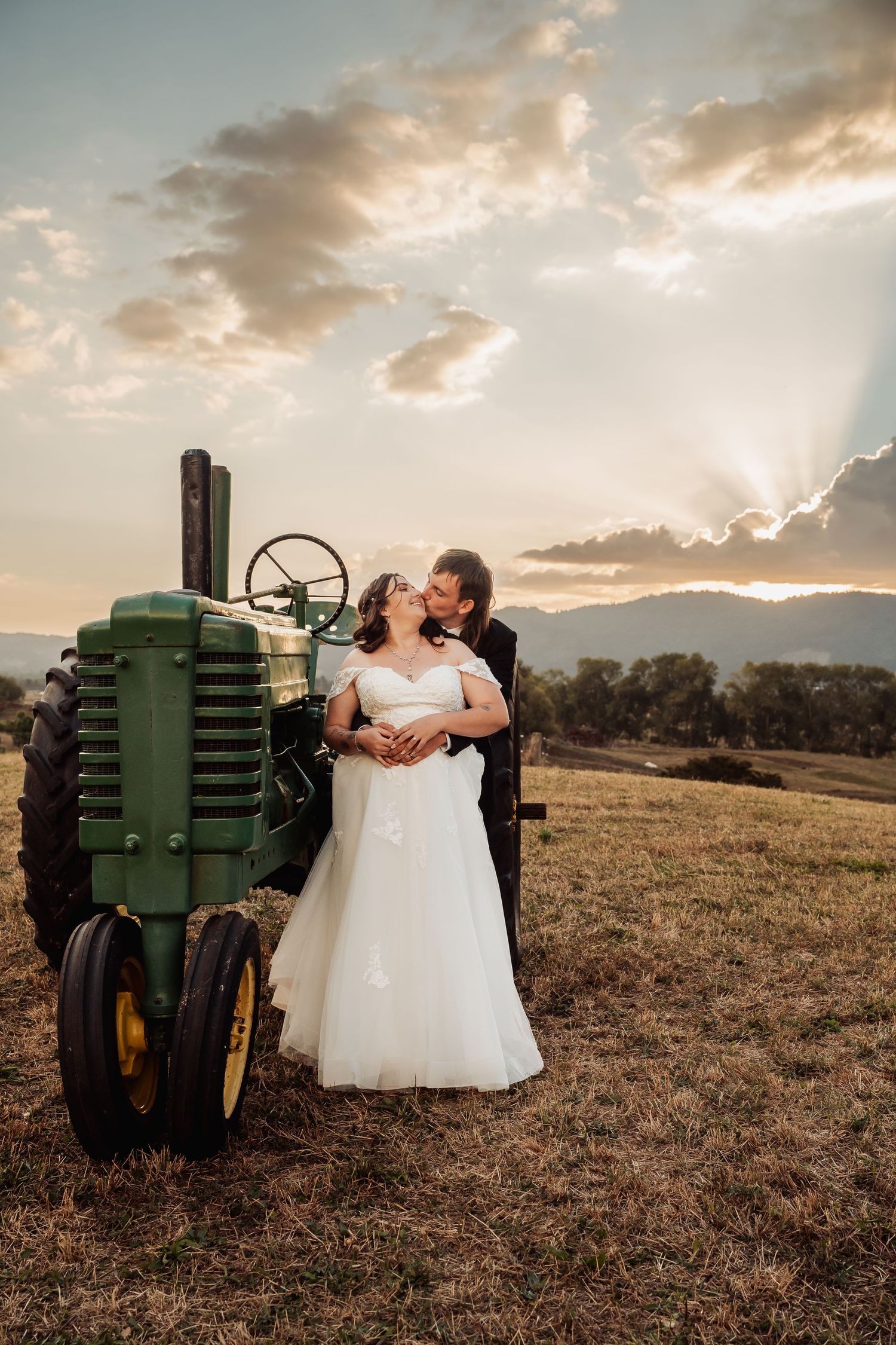 Bride and groom embrace beside a vintage green John Deere tractor at sunset on a rural farm property.