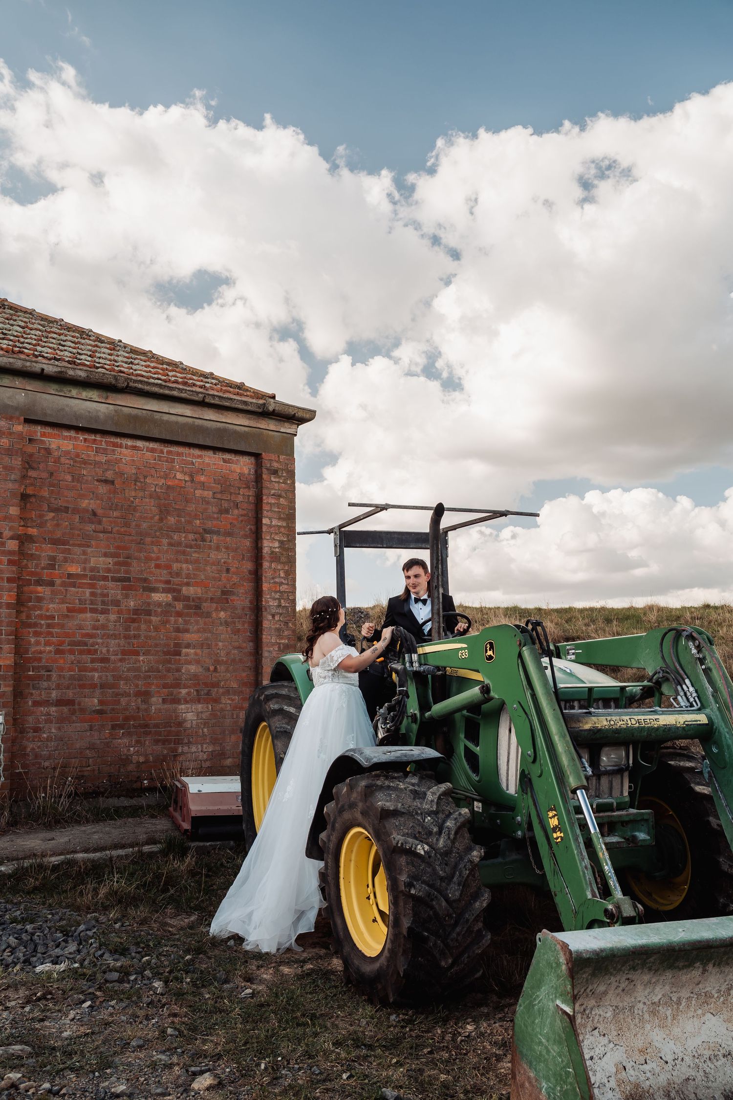 Bride and groom posing together on a green John Deere tractor beside a brick building under a cloudy sky.