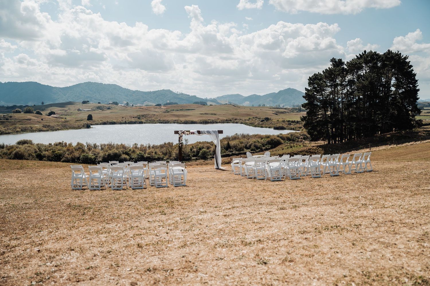 Outdoor wedding ceremony setup with white chairs facing lake and mountain backdrop on sunny day.