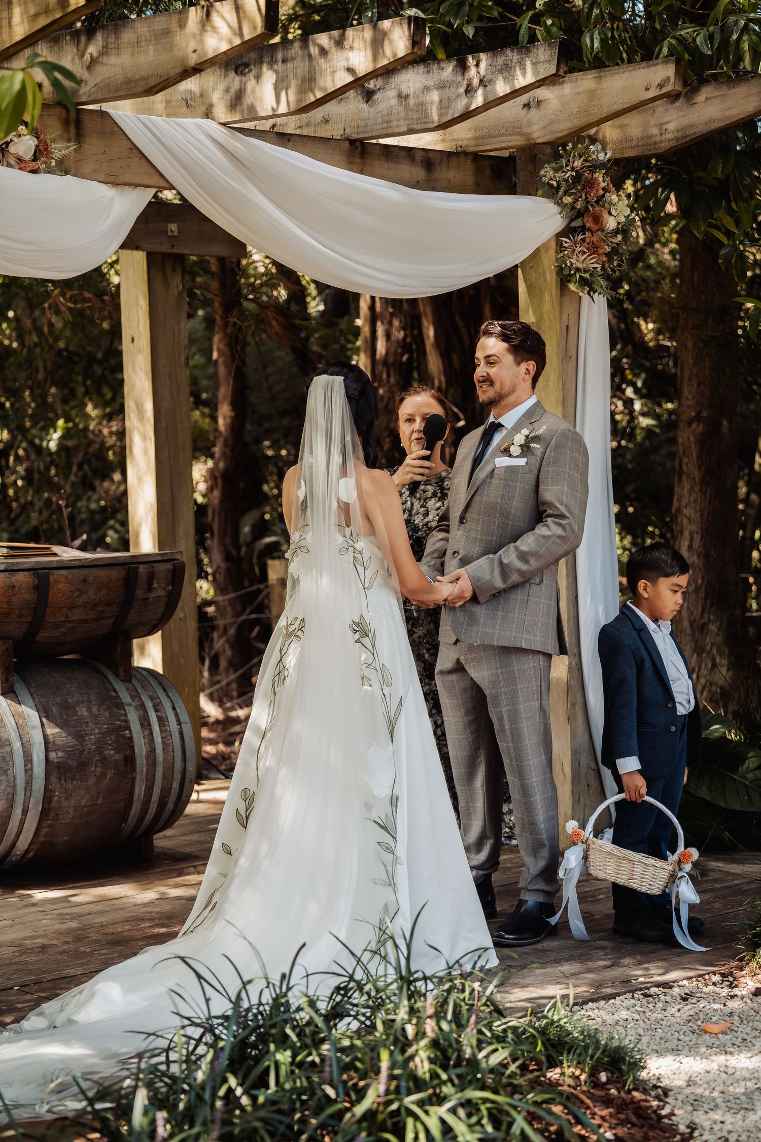 Bride and groom exchange vows under a draped wooden pergola at an outdoor garden wedding ceremony with ring bearer.