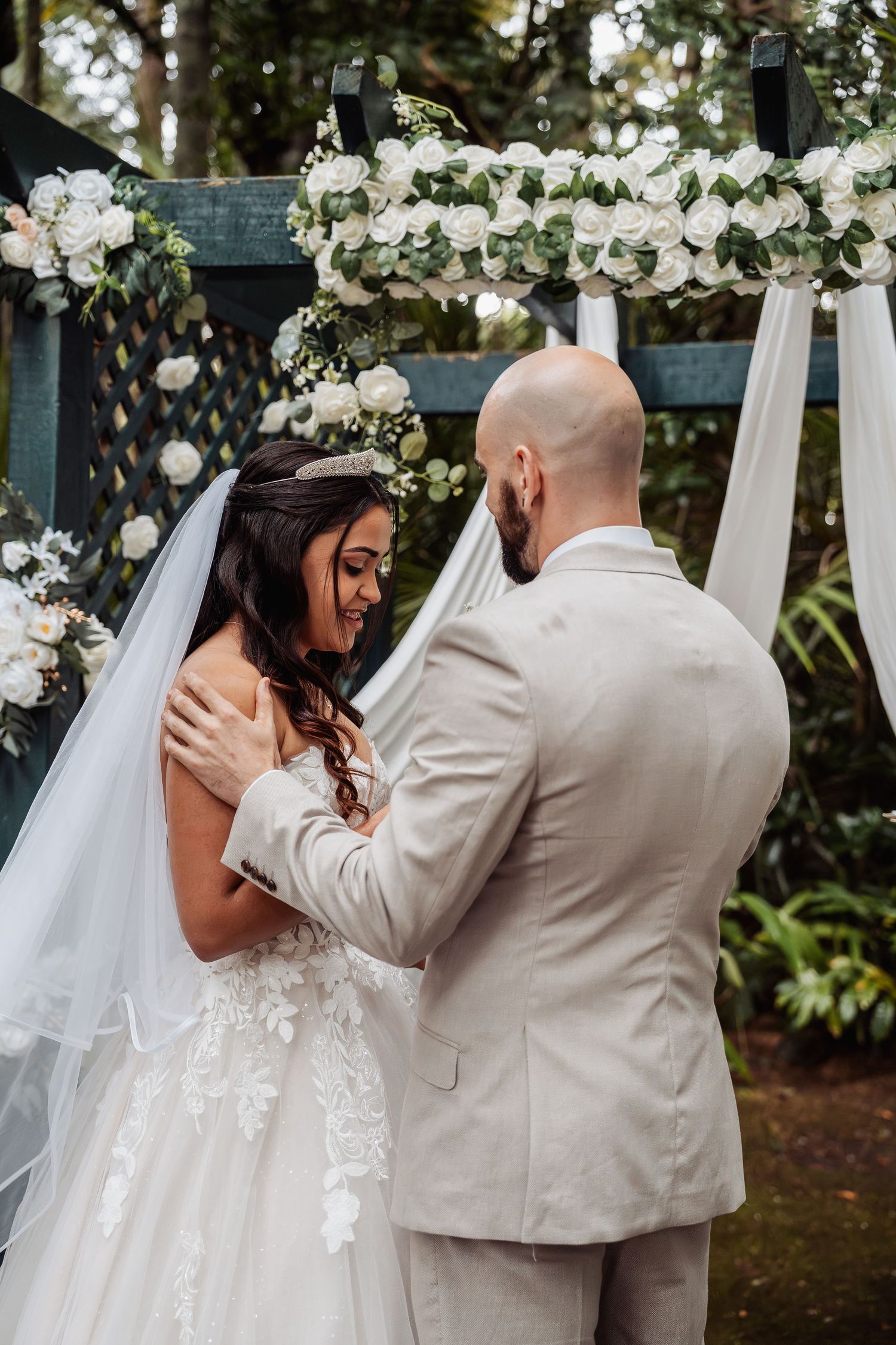 Bride and groom share a tender moment at outdoor wedding altar adorned with white roses and greenery.