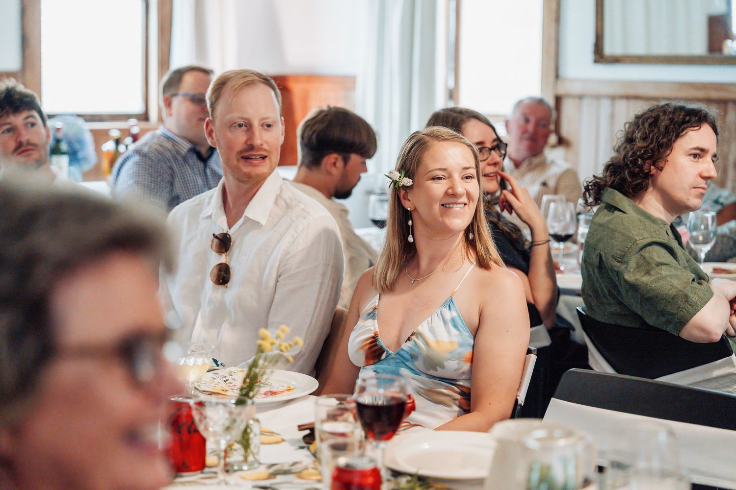 Wedding guests smiling and laughing at a reception dinner table with wine glasses and floral centerpieces.