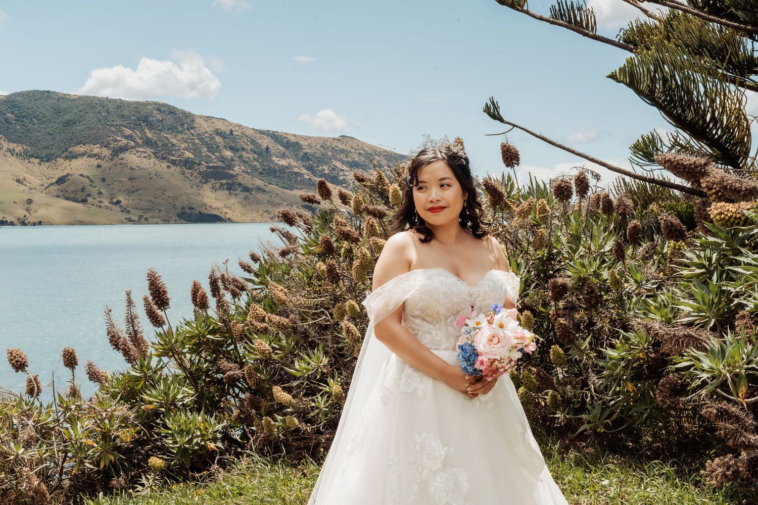 Bride in off-shoulder white gown holding colorful bouquet by scenic lake with lush green hills backdrop.