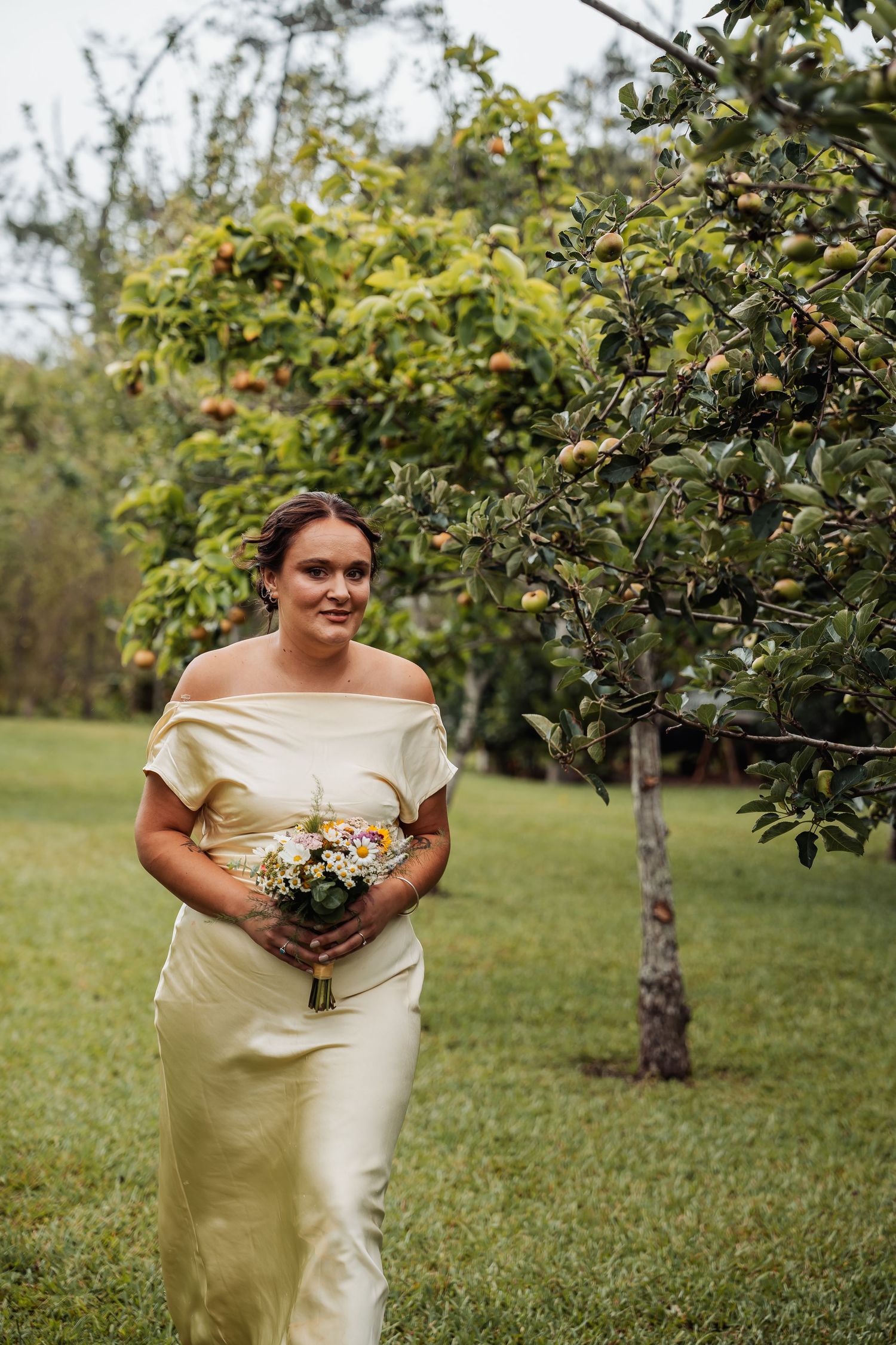 Bridesmaid in champagne satin off-shoulder gown holding a wildflower bouquet near apple trees.