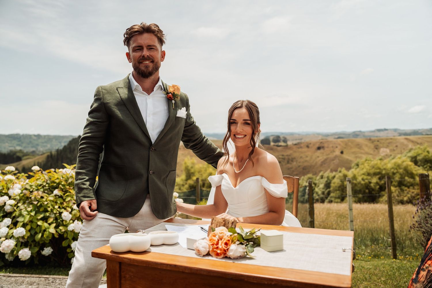 Smiling bride and groom sign wedding register at outdoor table with scenic countryside backdrop.