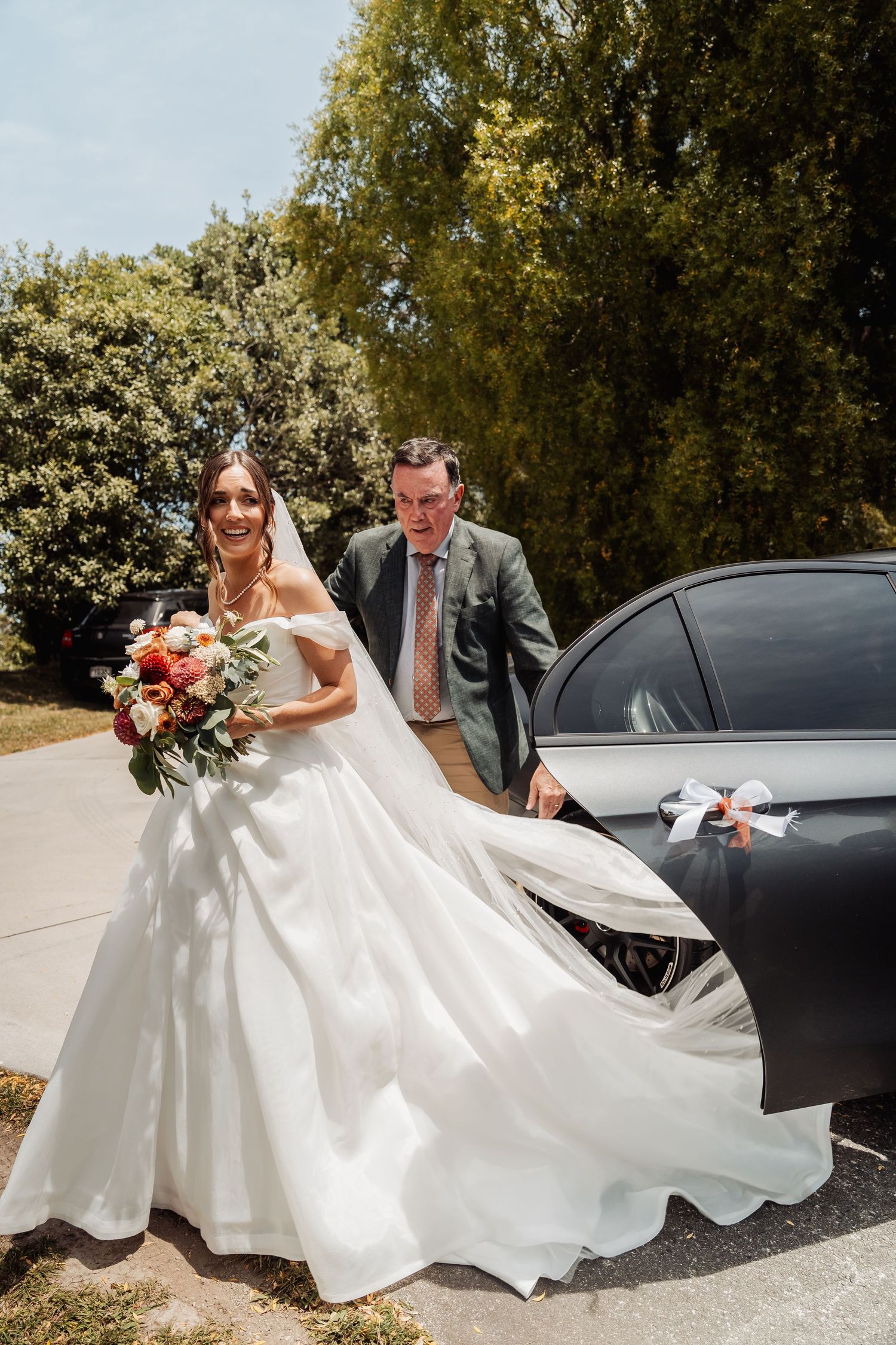Smiling bride in white gown holding colorful bouquet arriving at wedding venue with escort.
