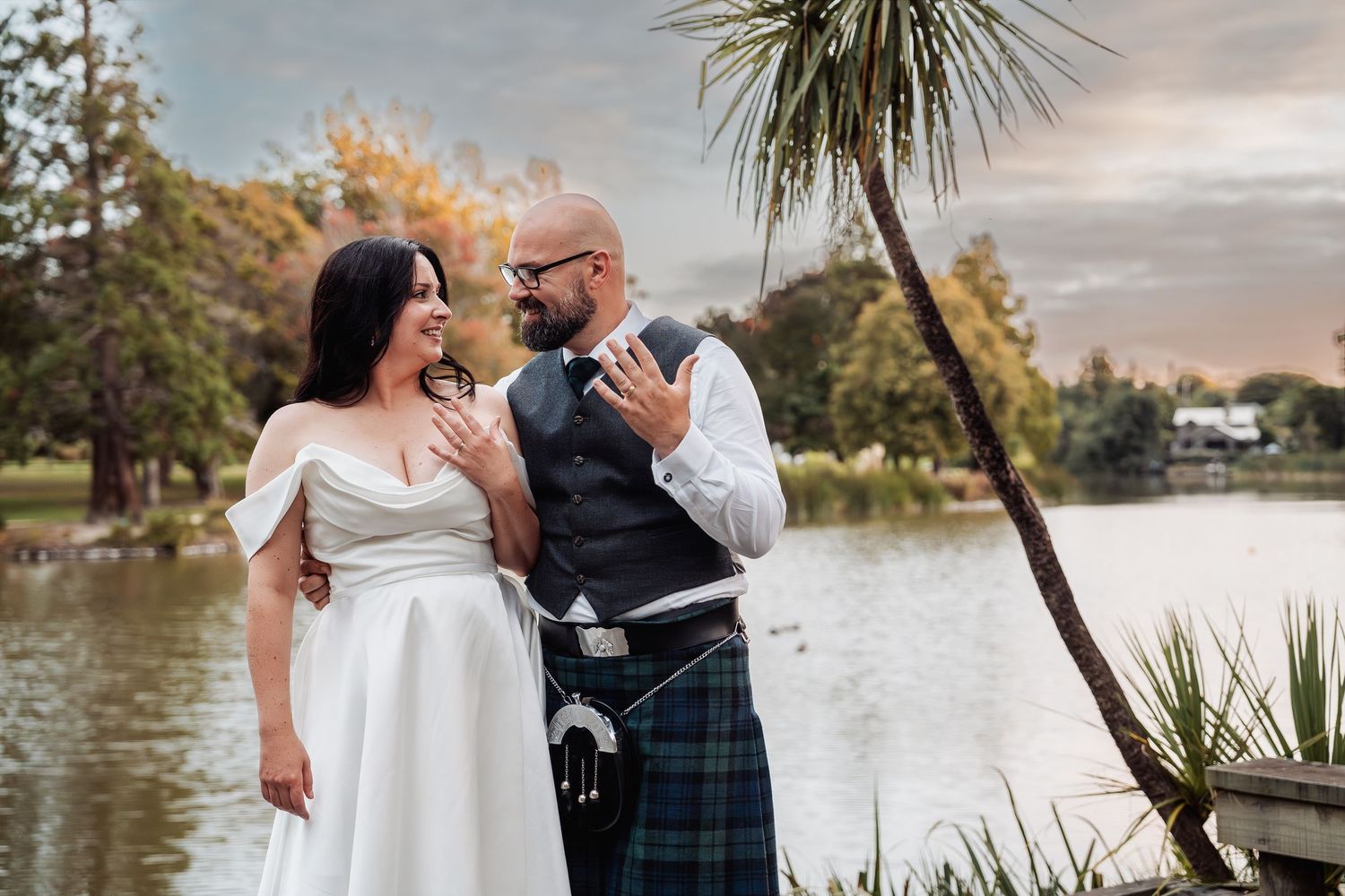 Bride and groom in Scottish kilt share a romantic moment beside a tranquil lake at golden hour.