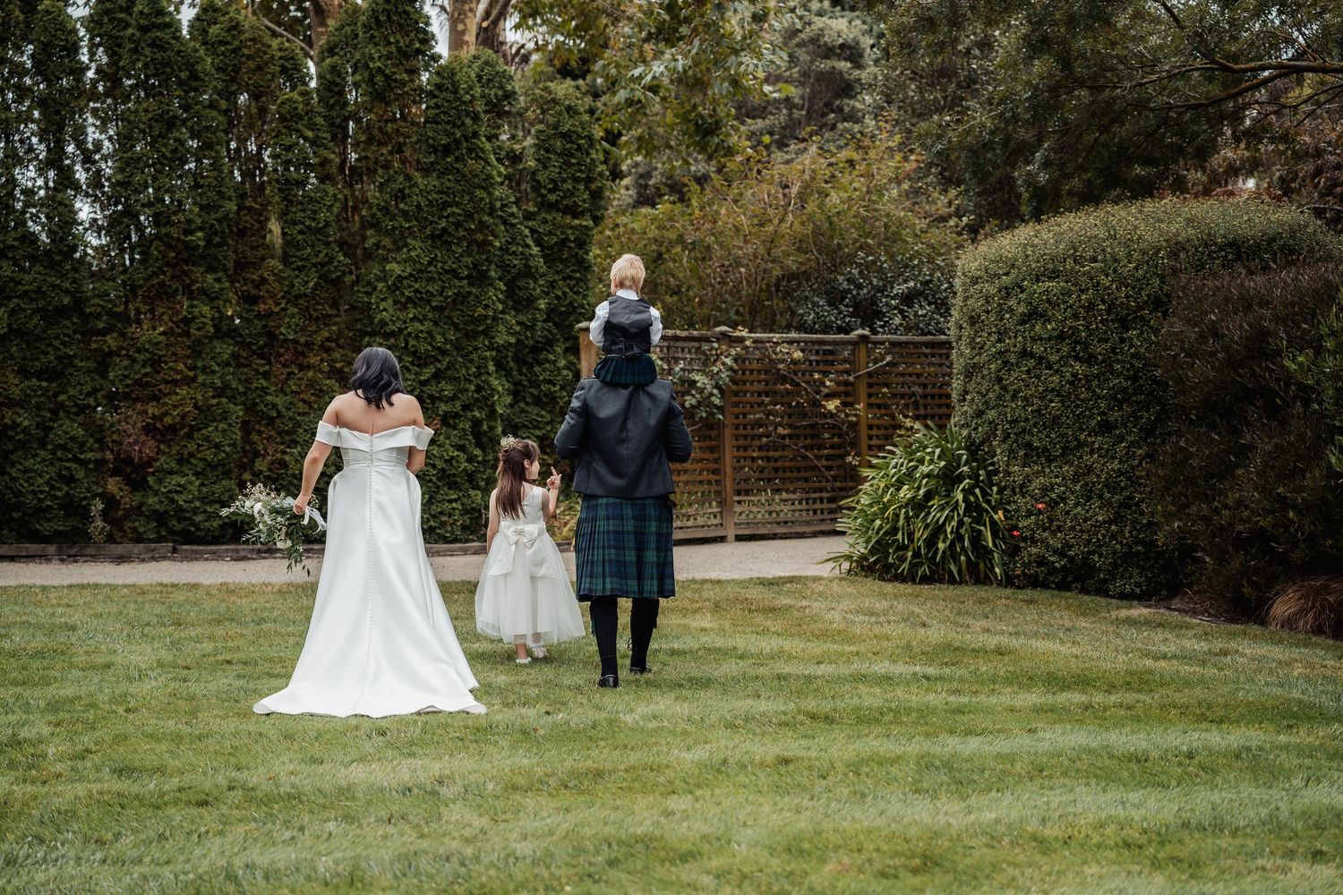 Wedding family portrait on green lawn with bride in white gown, groom in kilt holding child, and flower girl outdoors.