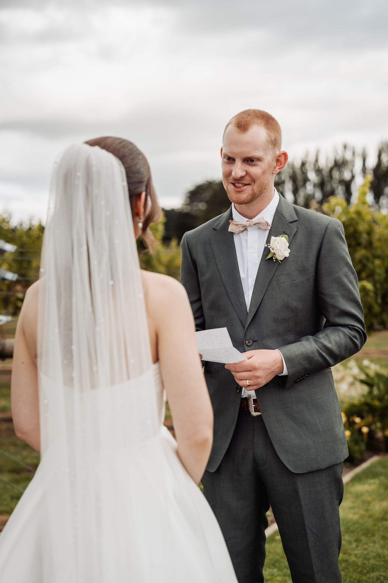 Groom reads vows from paper while smiling at veiled bride during outdoor vineyard wedding ceremony.
