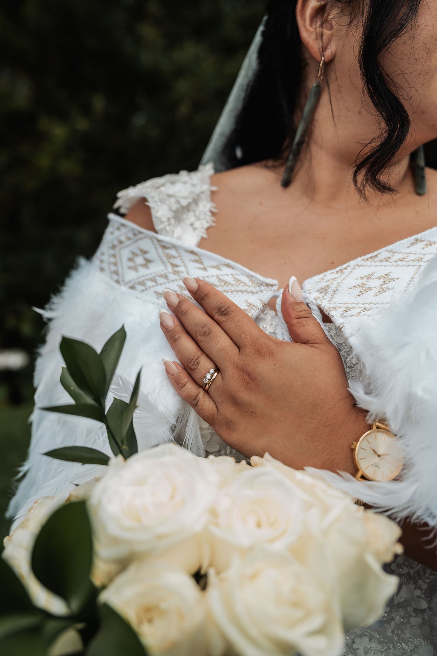 Close-up of bride holding white rose bouquet wearing wedding ring and gold watch in lace feather dress.