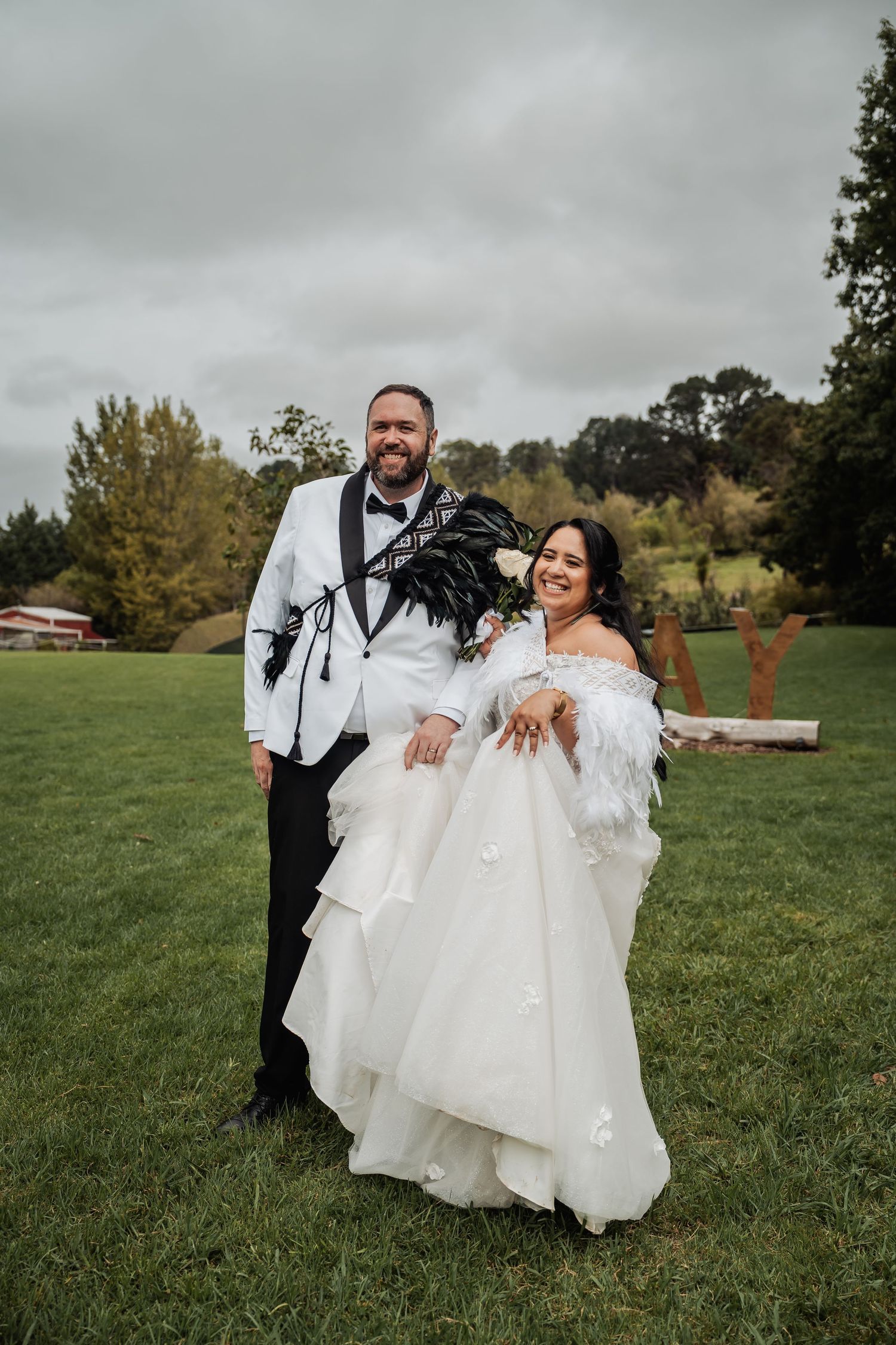Smiling bride and groom pose together on green lawn, bride in layered white gown with feather wrap.