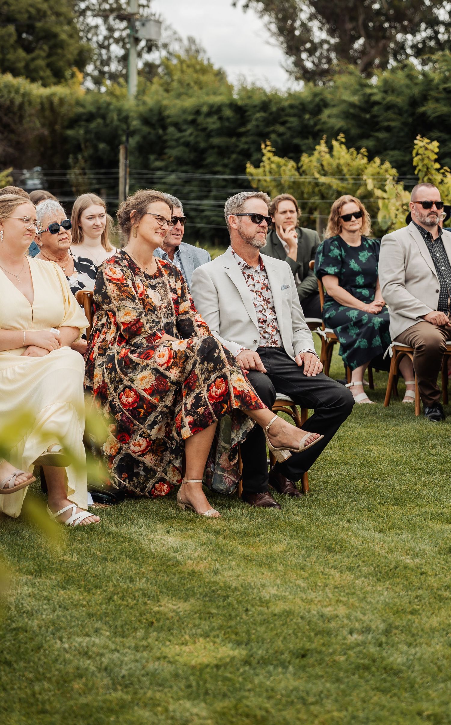 Wedding guests seated outdoors on grass enjoying a sunny garden ceremony.