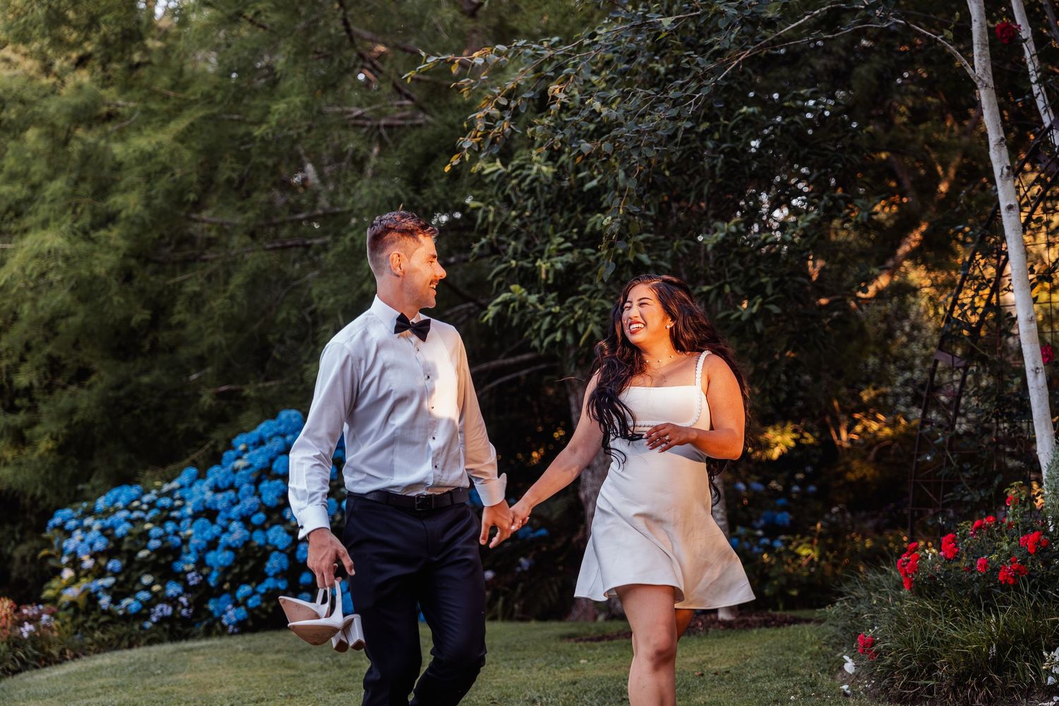 Newlywed couple holds hands walking through vibrant garden with colorful flowers during golden hour evening light.