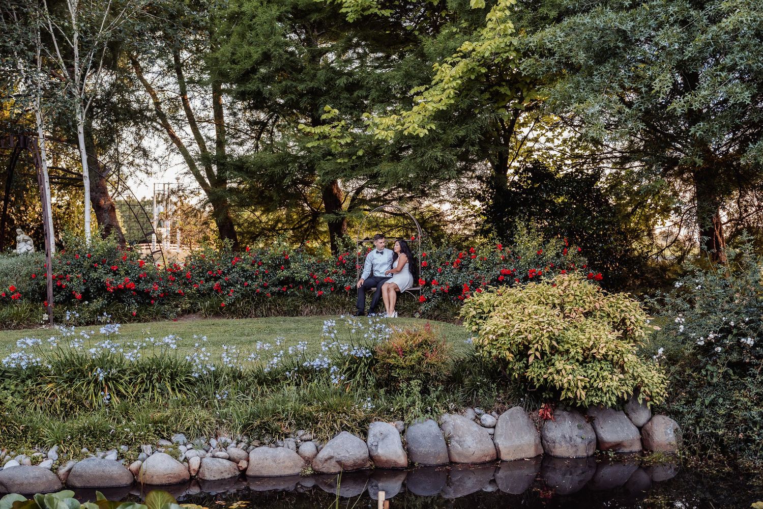 Bride and groom sitting together near a tranquil pond surrounded by colorful garden flowers.