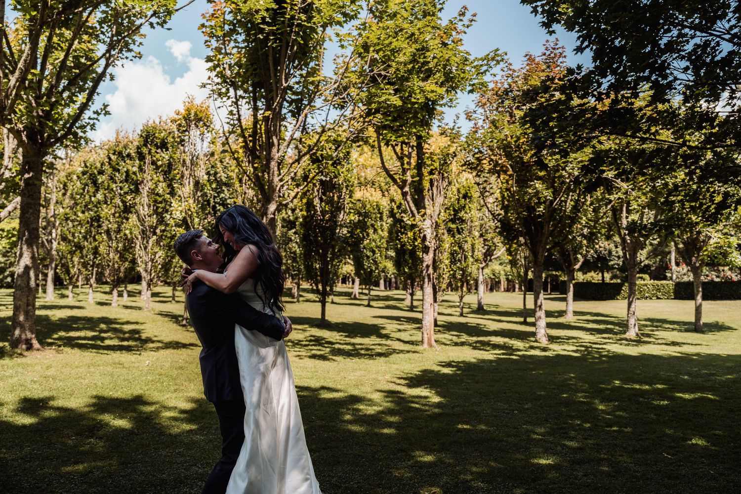 Groom lifts bride in white wedding dress in a romantic embrace surrounded by lush green trees outdoors.