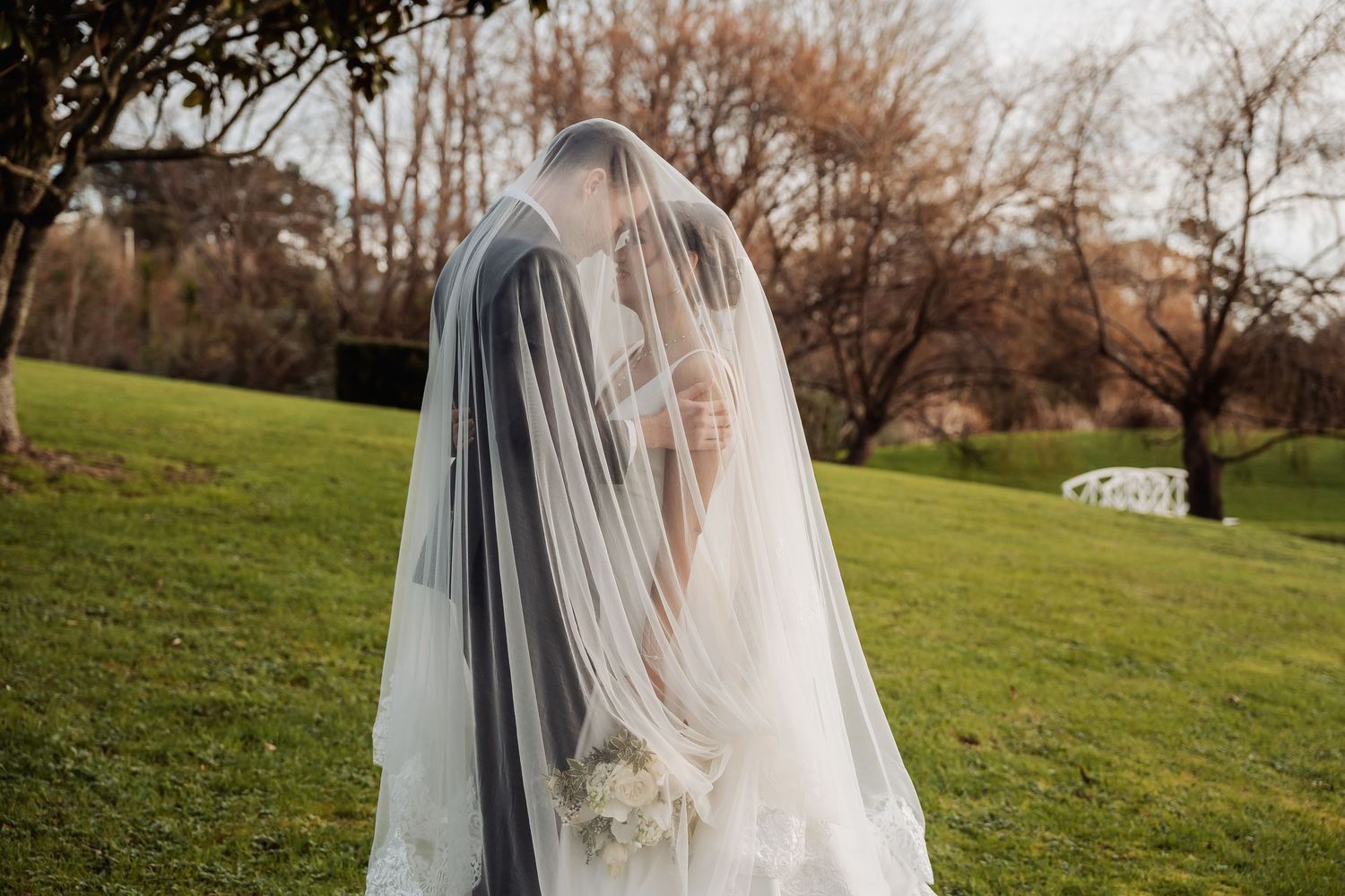 A long flowing wedding veil drapes over a couple on a grassy hillside.